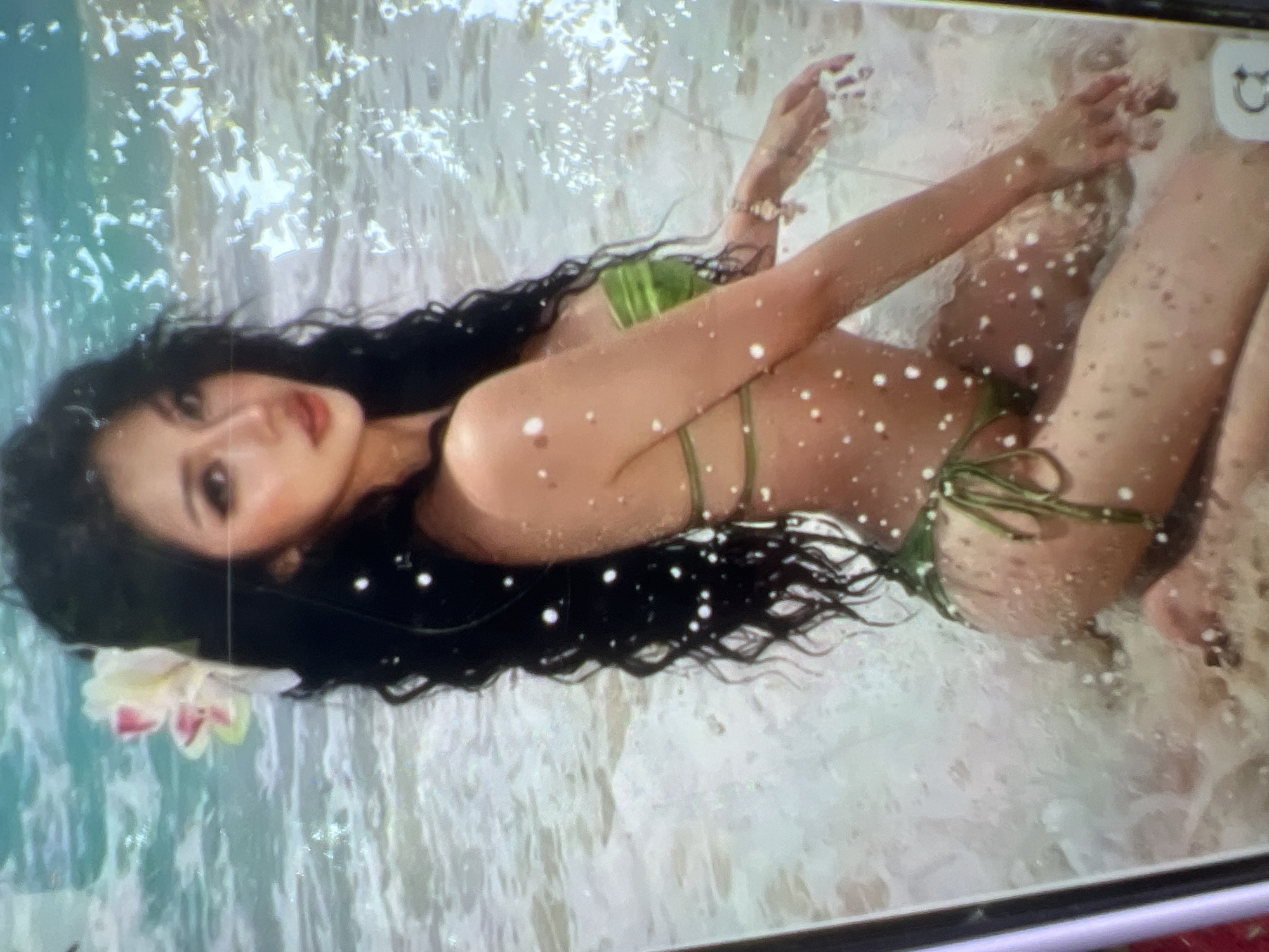 The image shows a young Asian woman with long, dark, wavy hair adorned with a white and pink flower, wearing a green bikini. She is kneeling in shallow ocean water on a sandy beach. The water is clear with white foam and ripples, and the sand is wet and speckled with small white dots, possibly sea spray or reflections. The woman is looking towards the viewer with a neutral expression. In the background, the ocean stretches out with turquoise and white water. There are no discernible buildings or structures from Kara-Dıykan, Kyrgyzstan, visible in the image. The scene depicts a typical beach setting.