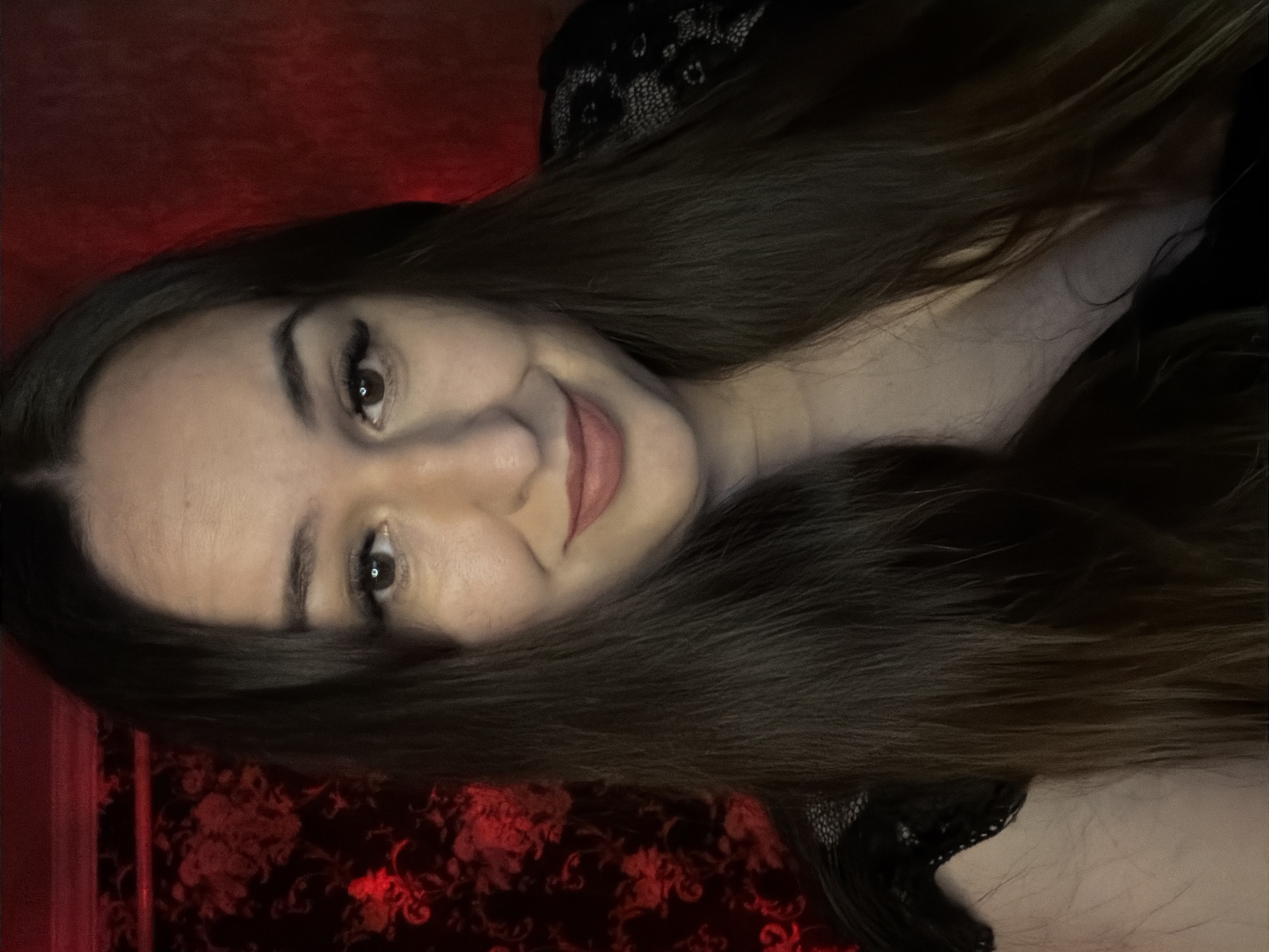 A medium close-up shot shows a woman with long, dark brown hair and brown eyes, looking directly at the camera with a slight smile. She is wearing dark clothing with lace detailing. The background consists of a dark, red patterned wallpaper on the left and a plain red background on the right. The lighting is dim and casts a reddish hue over the scene. There is no indication of the location being in a city.