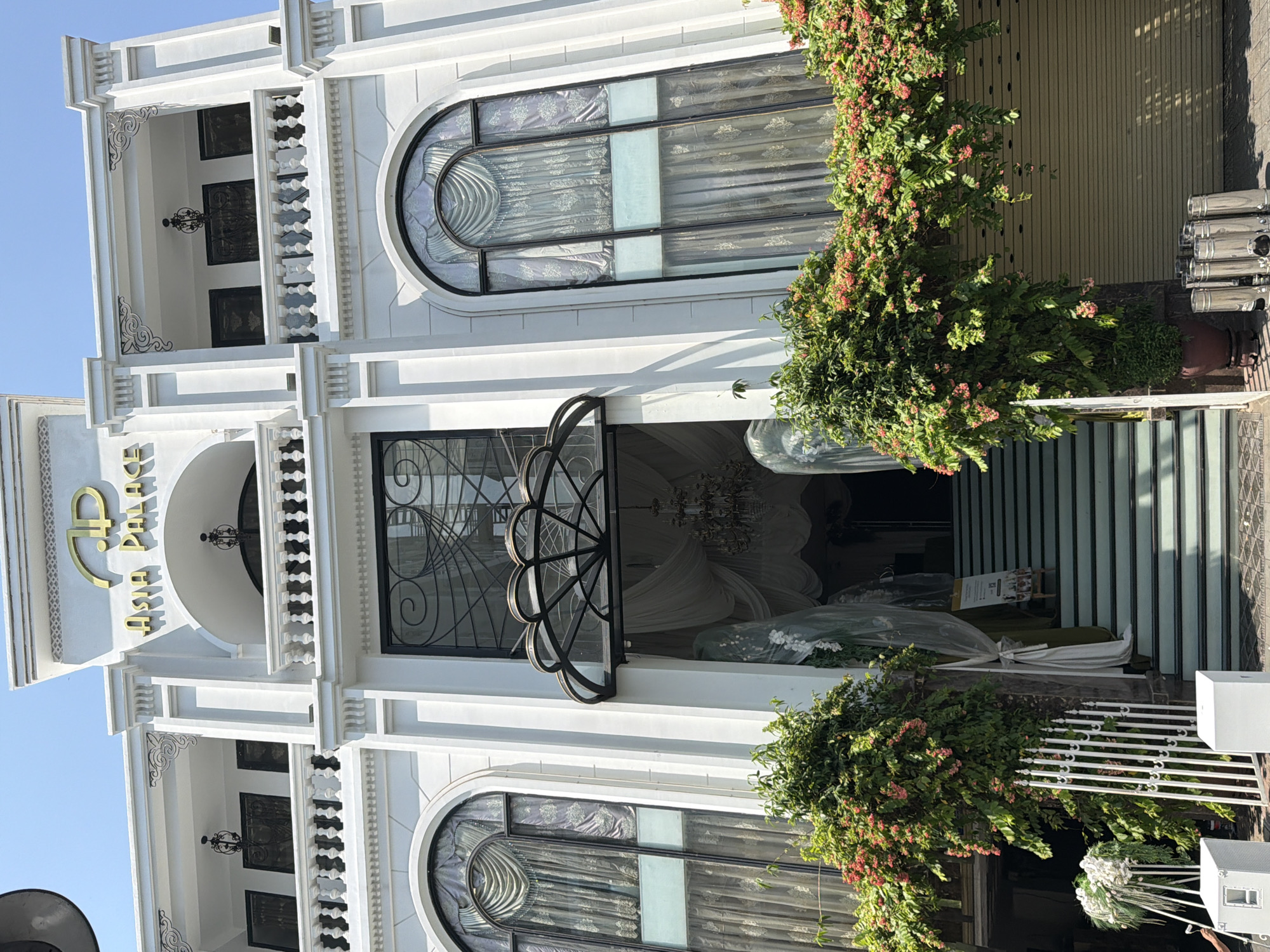 The image shows the ornate white facade of the Asia Palace venue, located in Thuận Hóa Ward, Vietnam. The building features multiple levels with decorative balconies, arched windows with elegant drapes, and intricate architectural details. Lush green foliage and flowering vines cascade over the lower portion of the building, framing the entrance. A set of stairs leads up to the main entrance, which is adorned with a decorative awning and appears to be draped with white fabric, suggesting a special event. White decorative metal fencing and large white cube-shaped objects, possibly speakers or equipment, are visible on the left side of the entrance, and several tall cylindrical metal objects are on the right. The sky above is clear and blue.