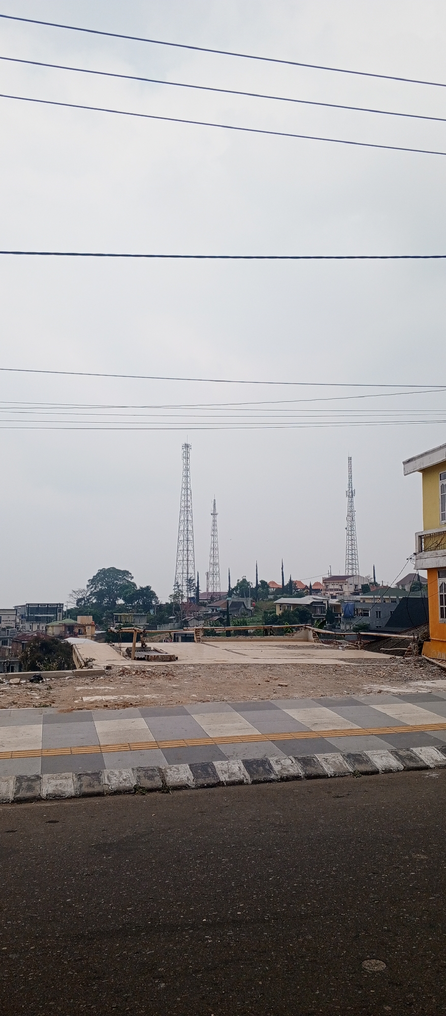 The image depicts a street scene in Sarangan, Indonesia, under an overcast sky. In the foreground, a paved road transitions into a sidewalk with a checkered pattern of grey and white tiles, bordered by a yellow tactile paving strip and a concrete curb. Beyond the sidewalk, a construction site is visible, featuring a concrete surface with debris and construction materials. A yellow excavator is parked on the site. In the mid-ground, a low-lying area shows signs of development with various buildings and structures. The background is dominated by several tall, lattice-like communication towers. Residential buildings, some with red-tiled roofs, are nestled on a hillside, interspersed with trees. A single-story building with a yellow facade and a white trim is partially visible on the right side of the image. Overhead, multiple power lines stretch across the frame. No people are visible in the image.