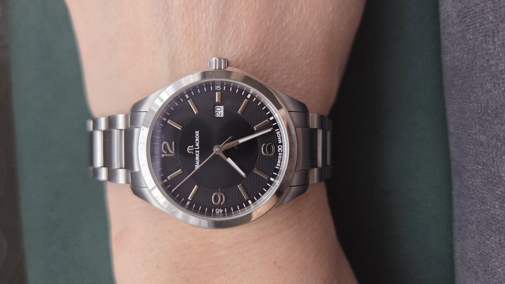 A close-up shot captures a person's wrist adorned with a Maurice Lacroix watch. The watch features a silver-toned metal bracelet with a brushed finish and a matching circular case. Its dark, possibly black, dial is vertically textured with subtle lines and displays gold-toned hour markers. Arabic numerals '12' and '6' are prominently visible, while other hours are marked with stick indices. A date window at the 3 o'clock position shows the number '20'. The watch hands are gold-toned and white, with the hour hand pointing just past '10' and the minute hand at '2', indicating approximately 10:10.
The watch crystal reflects light, and a small crown is visible on the right side of the case. The branding "MAURICE LACROIX" and the 'M' logo are clearly legible on the dial. Additional text, "SWISS 30 MADE", is visible near the 6 o'clock position, likely indicating "Swiss Made" and possibly "30M" for water resistance. The background is blurred, showing a dark green surface on the left and a darker, textured grey surface on the right, suggesting an indoor setting. The skin on the wrist is light-toned and shows natural texture.