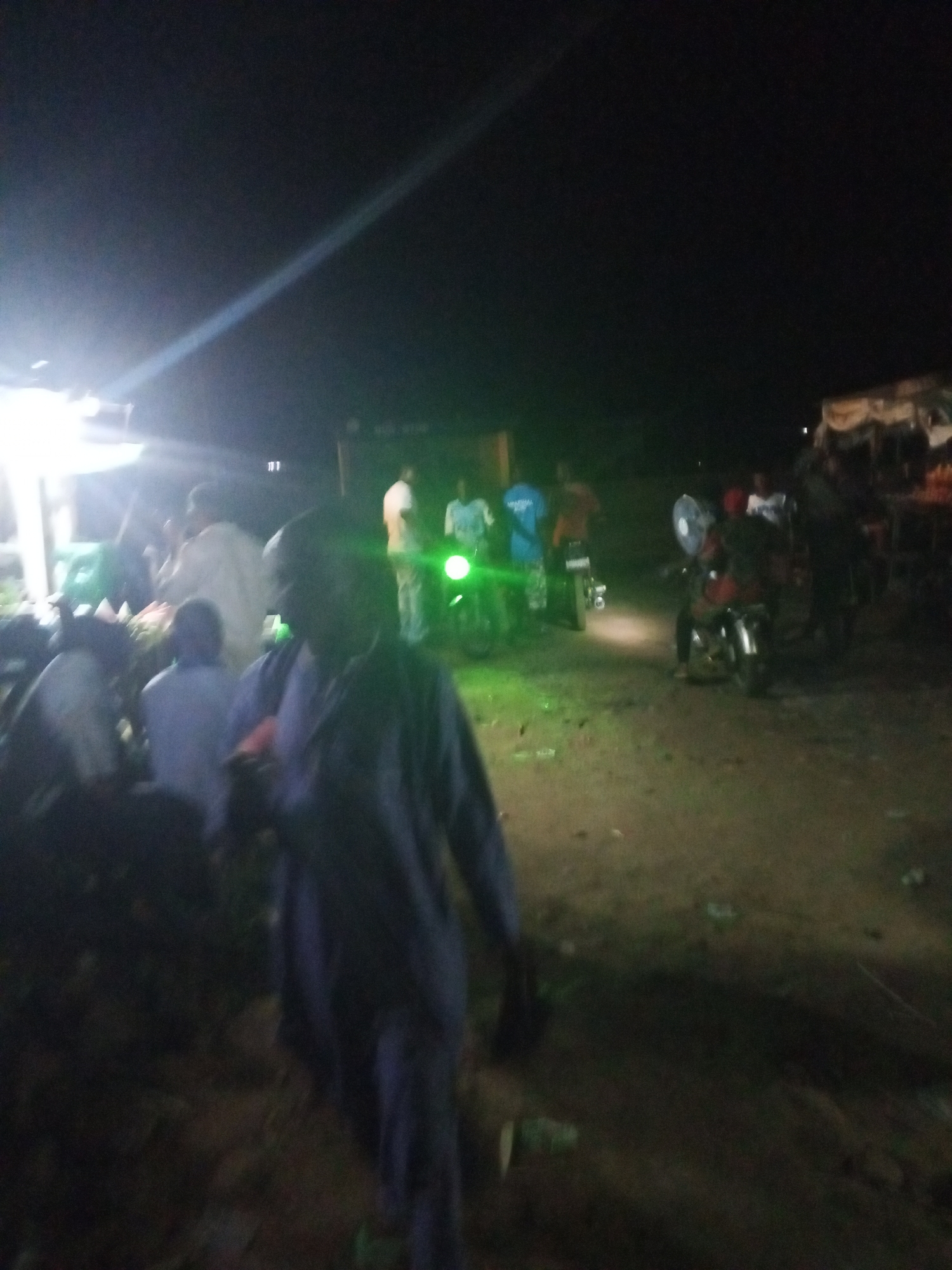 The image depicts an outdoor night scene in Lokoja, Nigeria, with a dimly lit area where people are gathered. Several individuals are standing and sitting near a light source that appears to be a market stall, where some produce is visible. Several motorcycles are parked nearby, some with their lights on, creating a source of illumination. The environment appears to be a market area or roadside with dirt or gravel ground. The overall scene shows a bustling but subdued environment with people likely engaged in commerce or gathering at the market location.