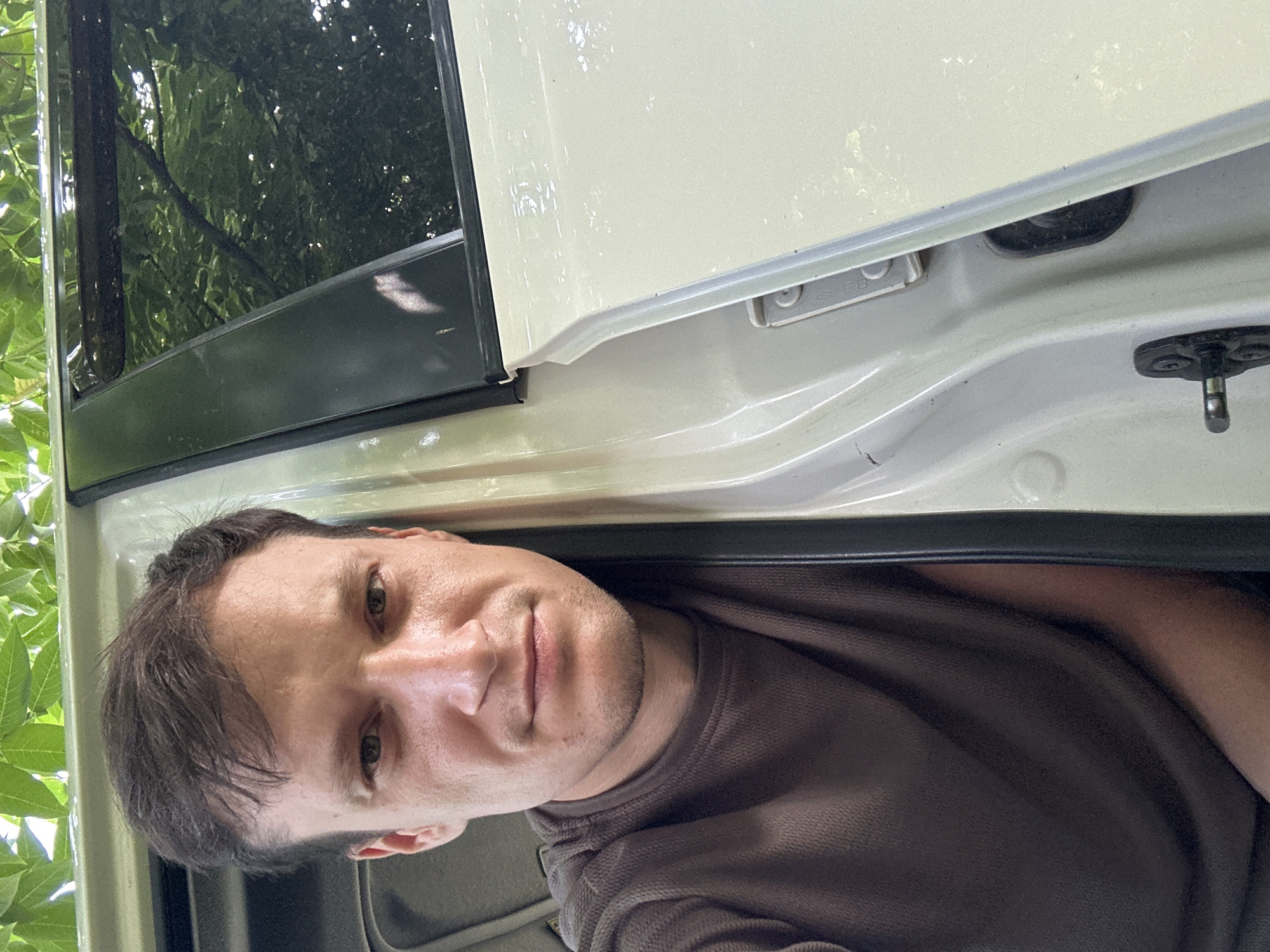 A person is positioned in the foreground, angled towards the camera, with their left side against the interior of a light-colored vehicle. The person has short, dark brown hair and is wearing a brown long-sleeved shirt. The vehicle's interior is visible, including a white door panel with visible hardware and the edge of a black window trim. Outside the vehicle, green foliage is visible, suggesting an outdoor environment. The image appears to be a selfie, taken at an eye-level perspective. The specific location in Makiv, Ukraine, is not determinable from the image alone.