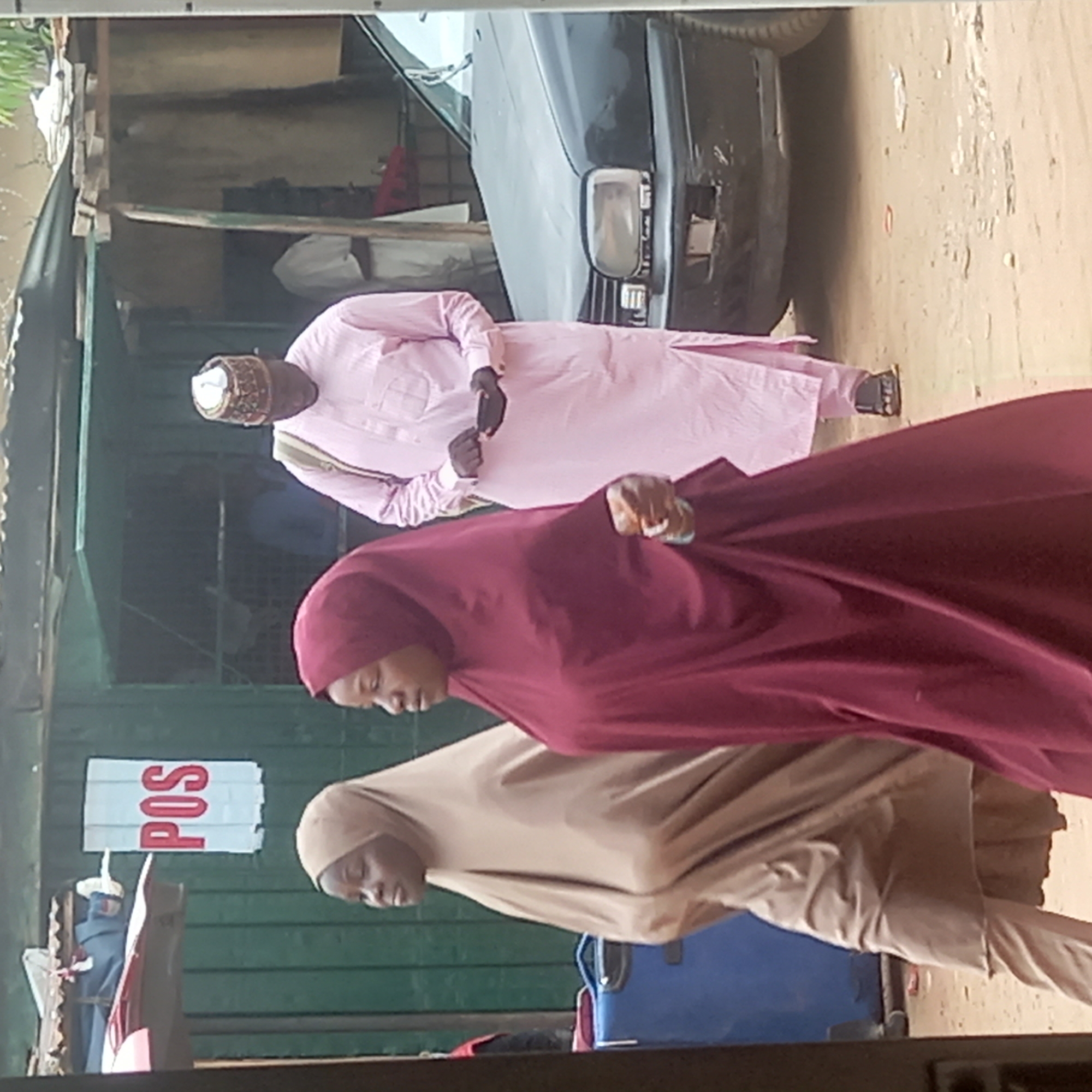 The image depicts three individuals in an outdoor setting. A man wearing a pink garment and a cap is followed by two women wearing headscarves. They are walking on a dirt road alongside a parked black car and a building with a "POS" (Point of Sale) sign. The location is consistent with an urban area in Dadi Riba, Nigeria.