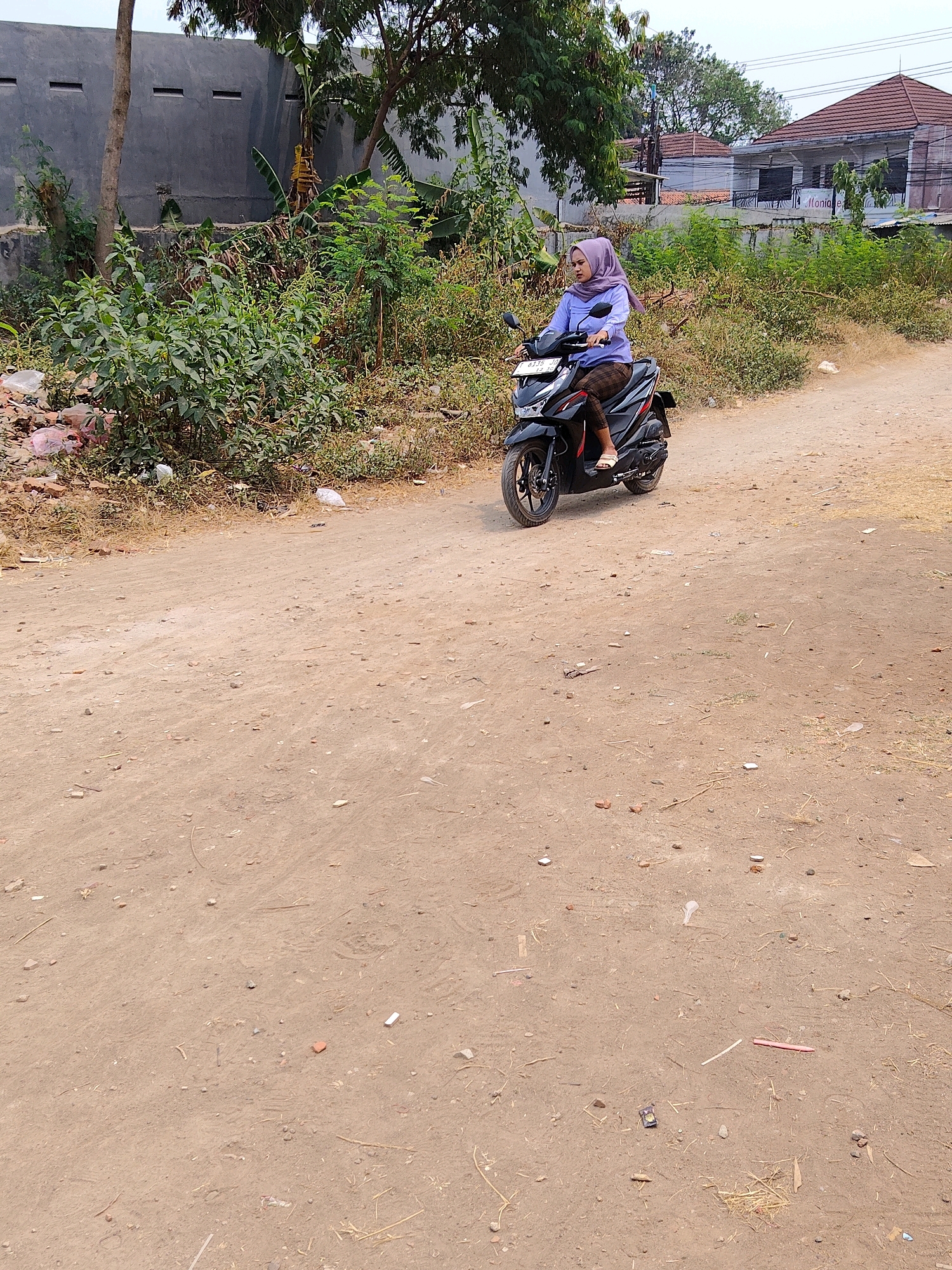 A young woman wearing a purple hijab and a light blue long-sleeved shirt is riding a black scooter on a dirt road. She is wearing patterned leggings and white sandals. The scooter has a license plate that reads "B 3336 JLL 12-25". The road is unpaved and appears to be in a rural or semi-urban area. Behind the woman, there is dense vegetation, a grey wall with horizontal openings, and a building with a tiled roof. To the right of the road, there are more buildings and trees. The scene is set in Purwakarta, Indonesia, likely on a sunny day.