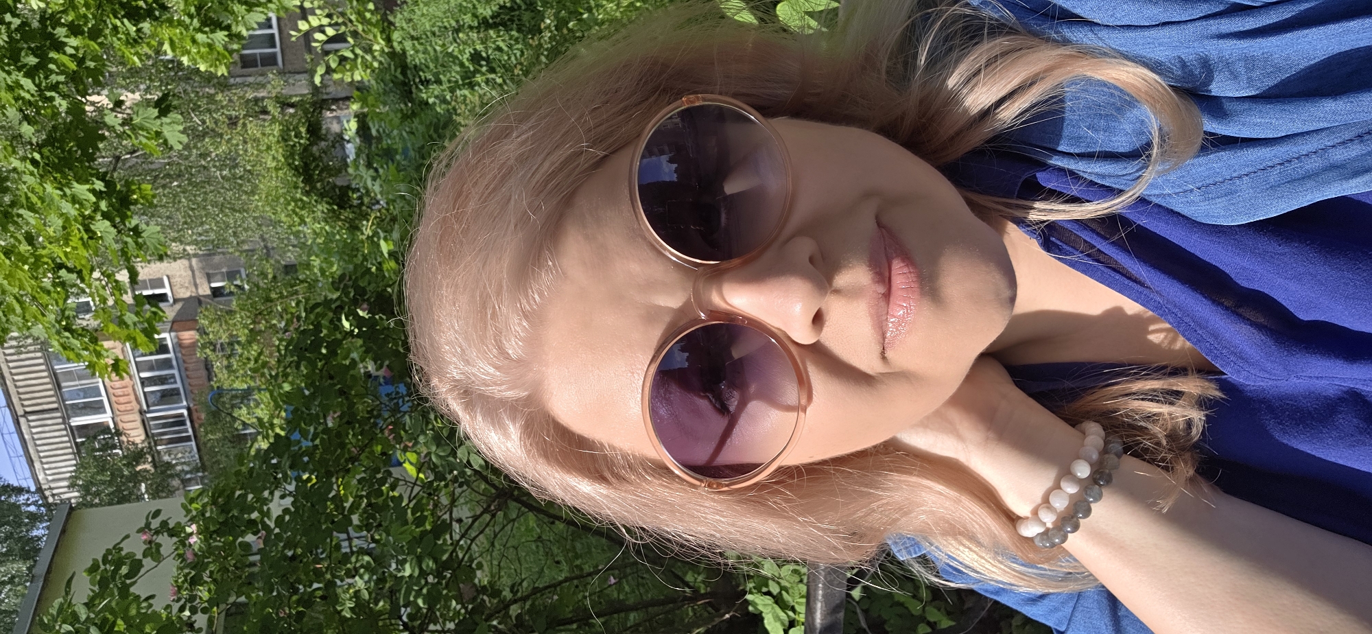 The image is a rotated close-up portrait of a person, likely a woman, with light blonde hair, captured outdoors in bright daylight. The subject's head is tilted, and the image is oriented so her head appears to be resting on its side. She is wearing large, round sunglasses with light pinkish frames, which reflect the bright sky and surrounding foliage. Her lips are slightly parted and have a glossy finish.
She is dressed in a blue garment, possibly a top or dress, which is visible draped around her upper body. On her left wrist, she wears two beaded bracelets: one composed of light, possibly pink or white, beads and another with darker, greyish beads.
The background is dominated by lush green foliage and trees, indicating an outdoor setting such as a garden, park, or a residential area with ample greenery. Parts of buildings are visible through the leaves, suggesting an urban or suburban environment. The strong sunlight creates clear highlights on her hair and skin, and distinct shadows among the leaves. The overall mood is casual and relaxed, indicative of a bright, sunny day. There is no visible text in the image.