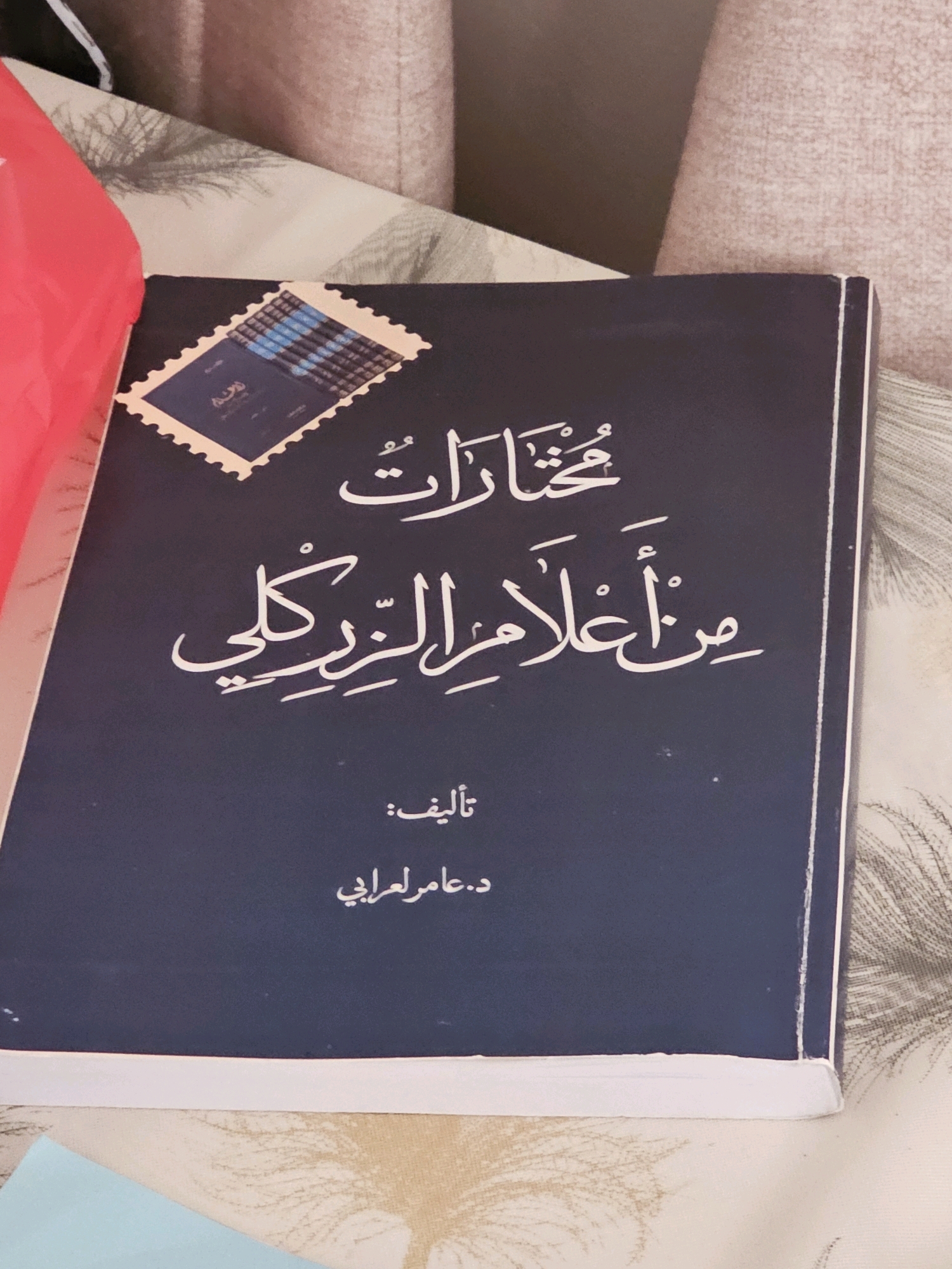 A dark blue book with white Arabic calligraphy lies on a surface with a textured pattern. The book is open, and the cover displays a postage stamp graphic in the upper left corner. The calligraphy on the cover translates to "Selections from the Scholars of the Century." Below this, it reads "Authored by: Dr. Amer Al-Arabi." The background suggests an indoor setting, possibly a living room or bedroom, with the edge of a red object visible on the left and part of a upholstered furniture item on the right. There is no information within the image that indicates a specific city or location.