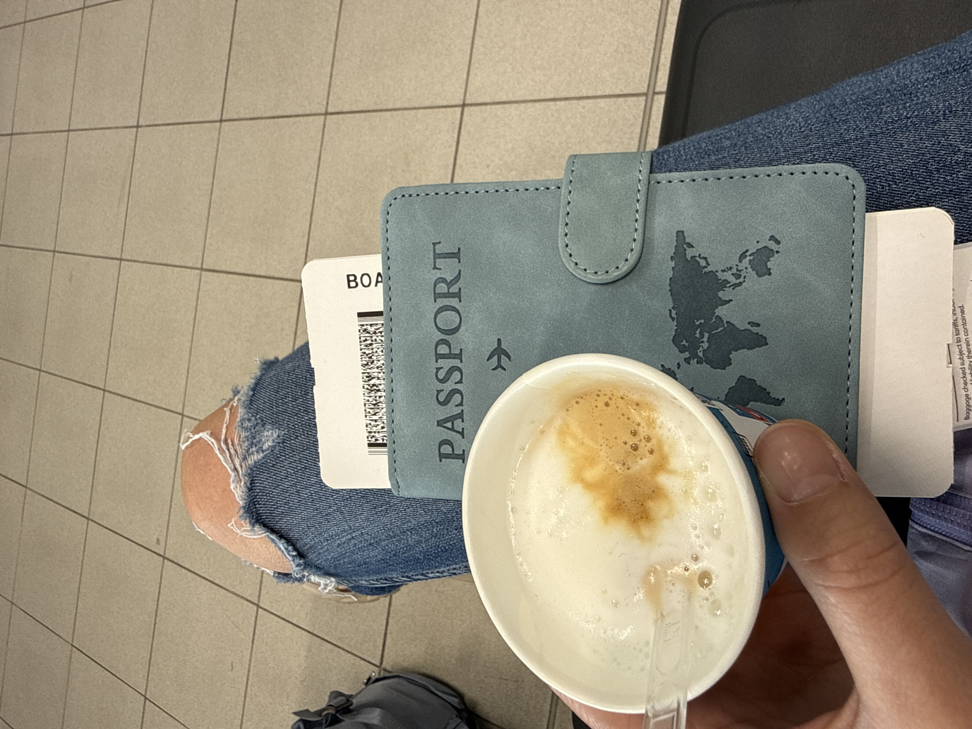 The image shows a person sitting in what appears to be an airport or a transit area with tiled flooring. The person is wearing blue jeans, which are ripped at the knee. On their lap, there is a blue passport holder with "PASSPORT" printed on it and a world map design. A boarding pass is tucked inside the passport holder. The person is holding a paper cup of coffee with foamed milk. The overall scene suggests the person is waiting to travel, possibly at an airport. It is not possible to determine the location in Sofia, Bulgaria, or even whether the photo was taken in Sofia based solely on the image.
