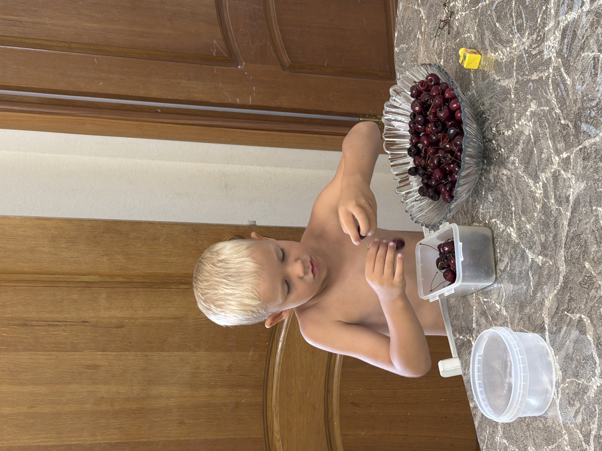 A young, shirtless boy with blonde hair is seated at a table, engaged in the act of pitting cherries. In front of him, there are three containers: a large glass bowl overflowing with fresh cherries, a clear plastic container filled with cherries, and an empty clear plastic container with a lid. The boy is holding a cherry in his right hand, and his left hand is positioned as if he is in the process of removing the pit. The table surface has a marbled pattern. In the background, wooden doors are visible. The overall scene appears to be indoors, in a domestic setting. No specific landmarks in Sochi, Russia, can be identified from the visual information provided.