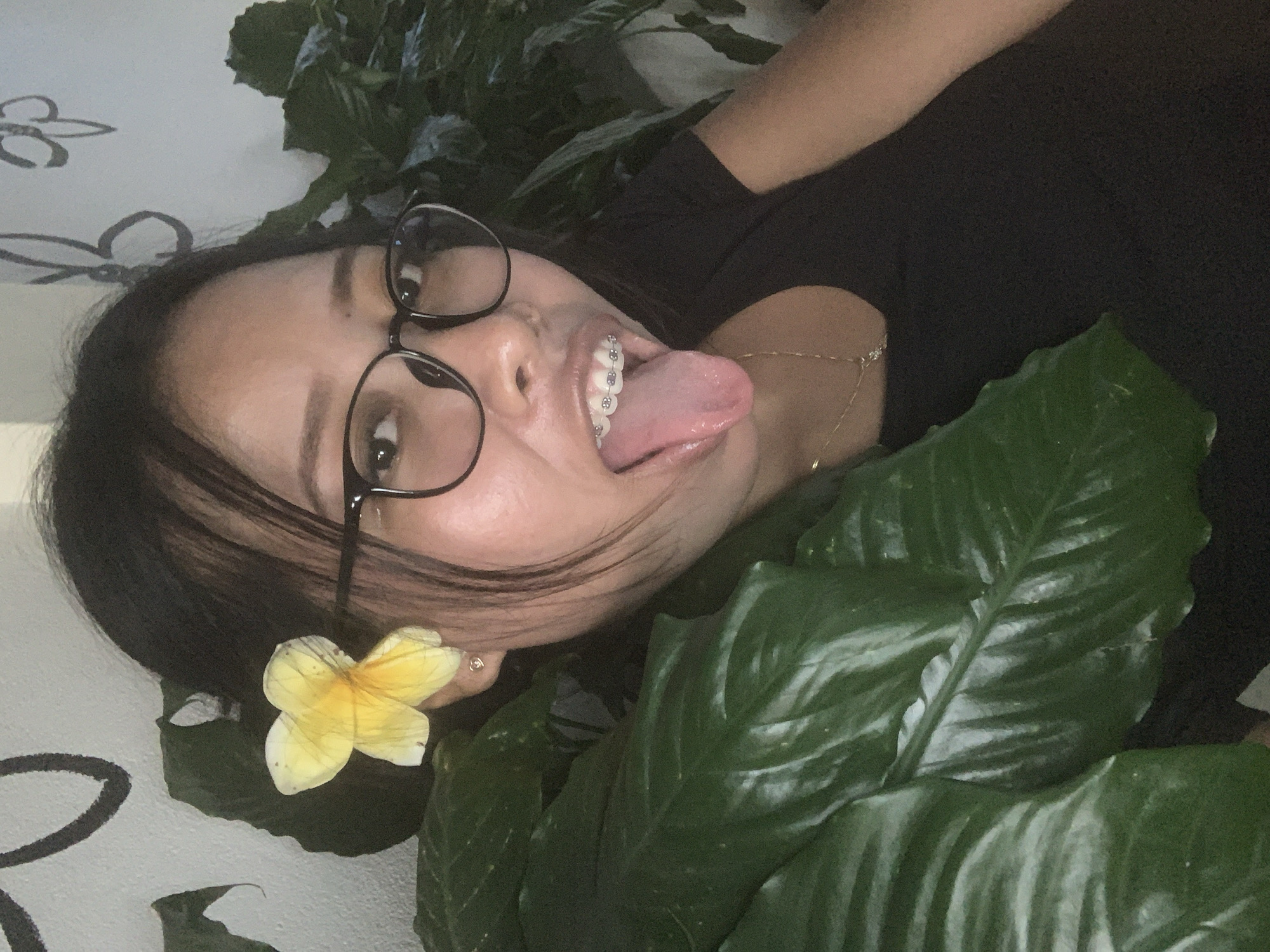 An informal portrait features a young woman playfully posing amidst lush green foliage, likely in the Ubud District, Indonesia. The image is oriented with the woman's head to the left, suggesting she is lying down and looking upwards, or the photo was taken at an angle.
The central subject is a woman with dark hair and a light complexion, wearing dark-rimmed glasses. She has clear dental braces on her upper teeth and is sticking her tongue out, conveying a mischievous or jovial expression. Her dark hair is pulled back, with some strands framing her face. A small, yellow plumeria flower is tucked behind her left ear, a common tropical bloom. She wears a black top or dress, and a delicate gold chain necklace is visible on her neck/shoulder.
She is surrounded by large, glossy, dark green leaves, suggesting she is nestled within dense plant life, possibly an indoor garden, patio area, or tropical foliage. Behind her, a light-colored wall with a decorative, dark floral or scroll-like pattern is visible, adding to the setting. The lighting is soft and indirect, indicating it could be daytime indoors or in a shaded outdoor area. No specific time of day or weather conditions are explicitly discernible, but the tropical flower and plants are consistent with the provided location context of Ubud. There is no visible text in the image.