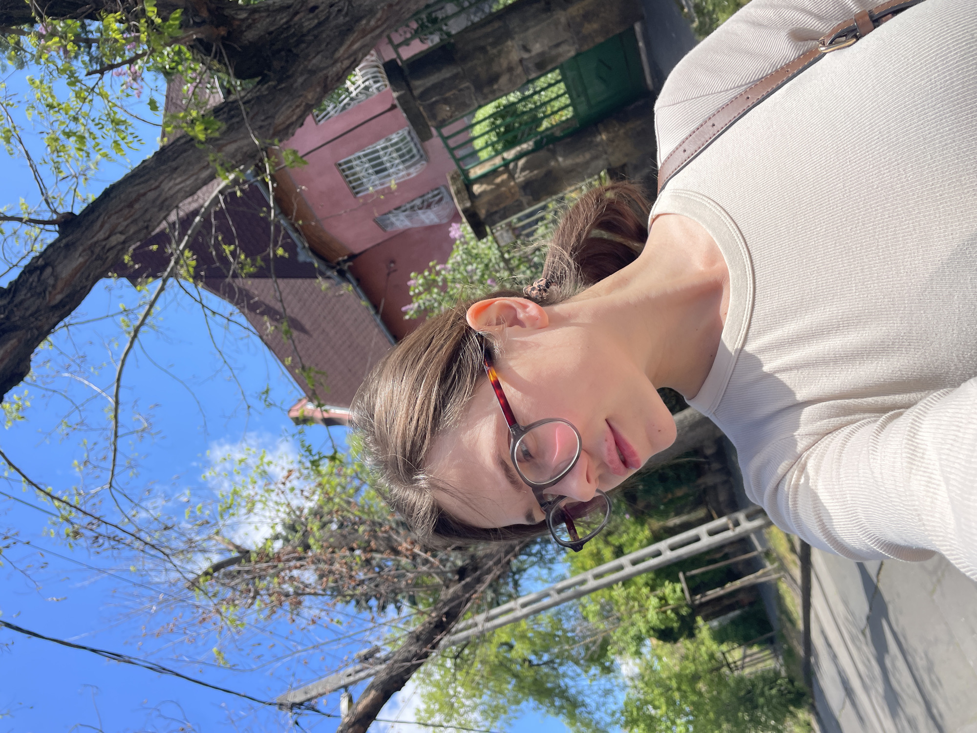 An upward-angled outdoor photograph, likely a selfie, captures a young woman against the backdrop of a sunny day in Budapest, Hungary.
The primary subject is a woman, visible from the chest up, with light brown hair pulled back. She wears circular glasses with tortoiseshell frames and a light-colored, possibly cream or off-white, ribbed long-sleeve top. A thin brown leather strap, indicative of a shoulder bag, diagonally crosses her chest. Her face is in profile, looking downwards with a neutral expression.
The scene depicts a bright, clear blue sky with sparse white clouds, suggesting a sunny day in spring or early summer, given the fresh, light green leaves on the numerous tree branches and trunks that frame the upper and left parts of the image. In the background, a distinctive multi-story building with a pink or peach facade and white-framed windows stands out. A dark green gate or door is visible near the building's base, above what appears to be a stone wall or foundation. A paved surface, possibly a street or path, and a concrete railing are partially visible in the lower right, hinting at an urban setting. No visible text is present.