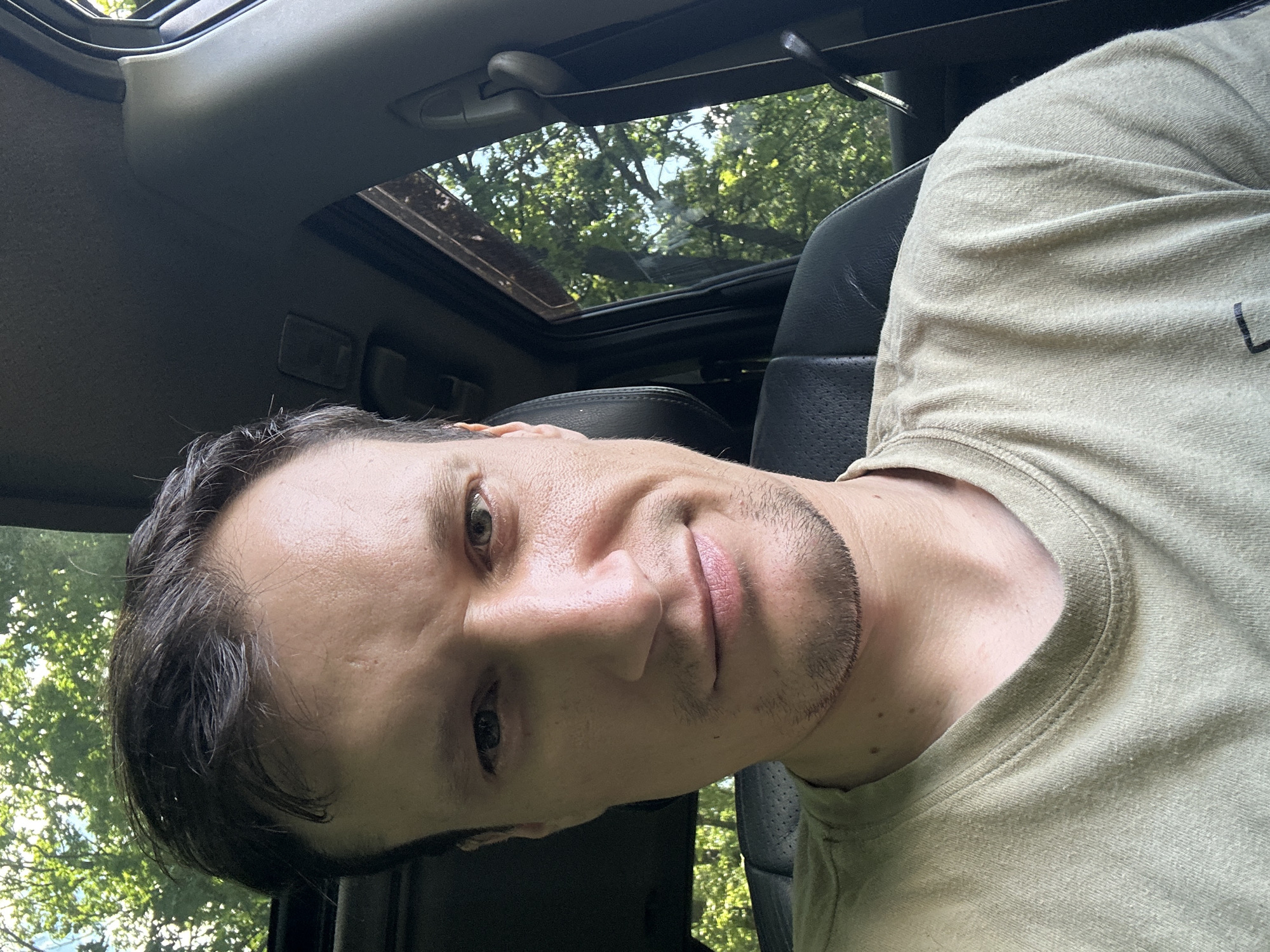 A young man is pictured inside a vehicle, looking at the camera. The man has short, dark hair, a light complexion, and appears to be in his late 20s or early 30s. He is wearing a light-colored t-shirt. The interior of the vehicle is visible, including a black leather seat and the ceiling. Outside the car, green trees and foliage are visible through the windows, suggesting an outdoor, possibly rural or park-like setting. There are no other people in the image. The lighting is natural and appears to be daylight. The image quality is good, with clear focus on the man's face. There is no information in the image that can confirm or deny the location as Shatava, Ukraine.