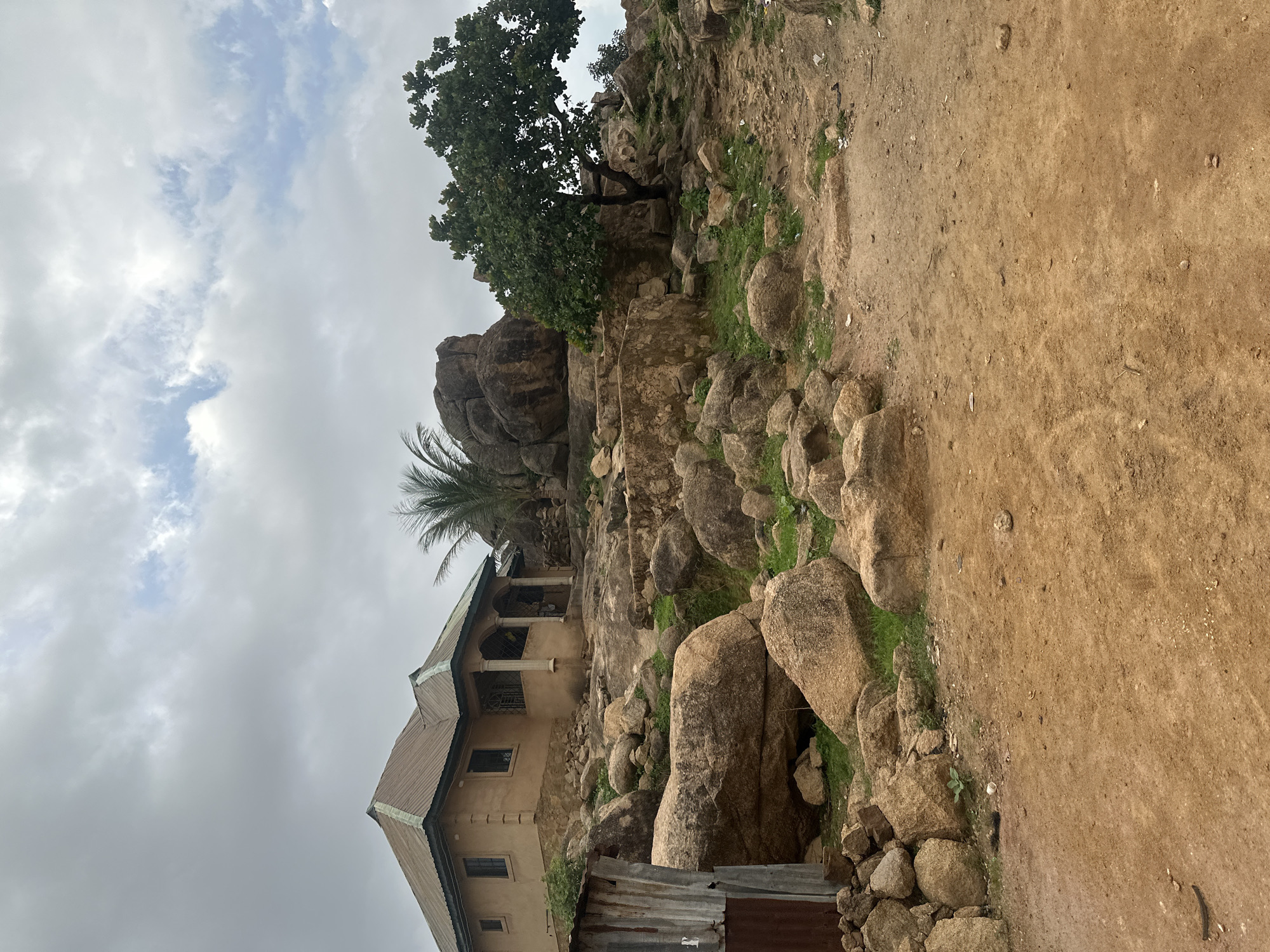 This image presents a rugged landscape dominated by large rock formations, characteristic of the granite outcrops found in places like Jos, Nigeria. The scene unfolds under an overcast or partly cloudy sky, indicating daytime with diffused light.
On the left side of the frame, a single-story house is visibly integrated into the rocky terrain. The house has a light brown or tan exterior and a sloped, dark-colored corrugated metal roof. A covered porch, supported by pillars, extends from the front of the house, offering a shaded outdoor space. Several windows are visible on its facade. A slender palm tree grows adjacent to the house, its fronds reaching above the roofline. At the very bottom left, a small structure or barrier constructed from weathered, possibly rusty, corrugated metal sheets is partially visible.
The central and right portions of the image are densely packed with numerous large, smooth, reddish-brown boulders of varying sizes, creating a natural, uneven landscape. Patches of green vegetation, including grass and small shrubs, cling to the soil in the crevices and between the rocks, adding touches of life to the stony environment. Further up and to the right, a larger tree with dense, leafy green foliage stands prominently among the rocks.
To the far right, a wide, unpaved area or dirt path is visible. This ground is a dry, light brown, earthy tone, with small stones and debris scattered across its surface, sloping gently upwards.
There are no people or specific activities depicted in the image. No visible text is present. The overall impression is one of human habitation thoughtfully adapted to and harmoniously coexisting with a dramatic, natural rocky environment.