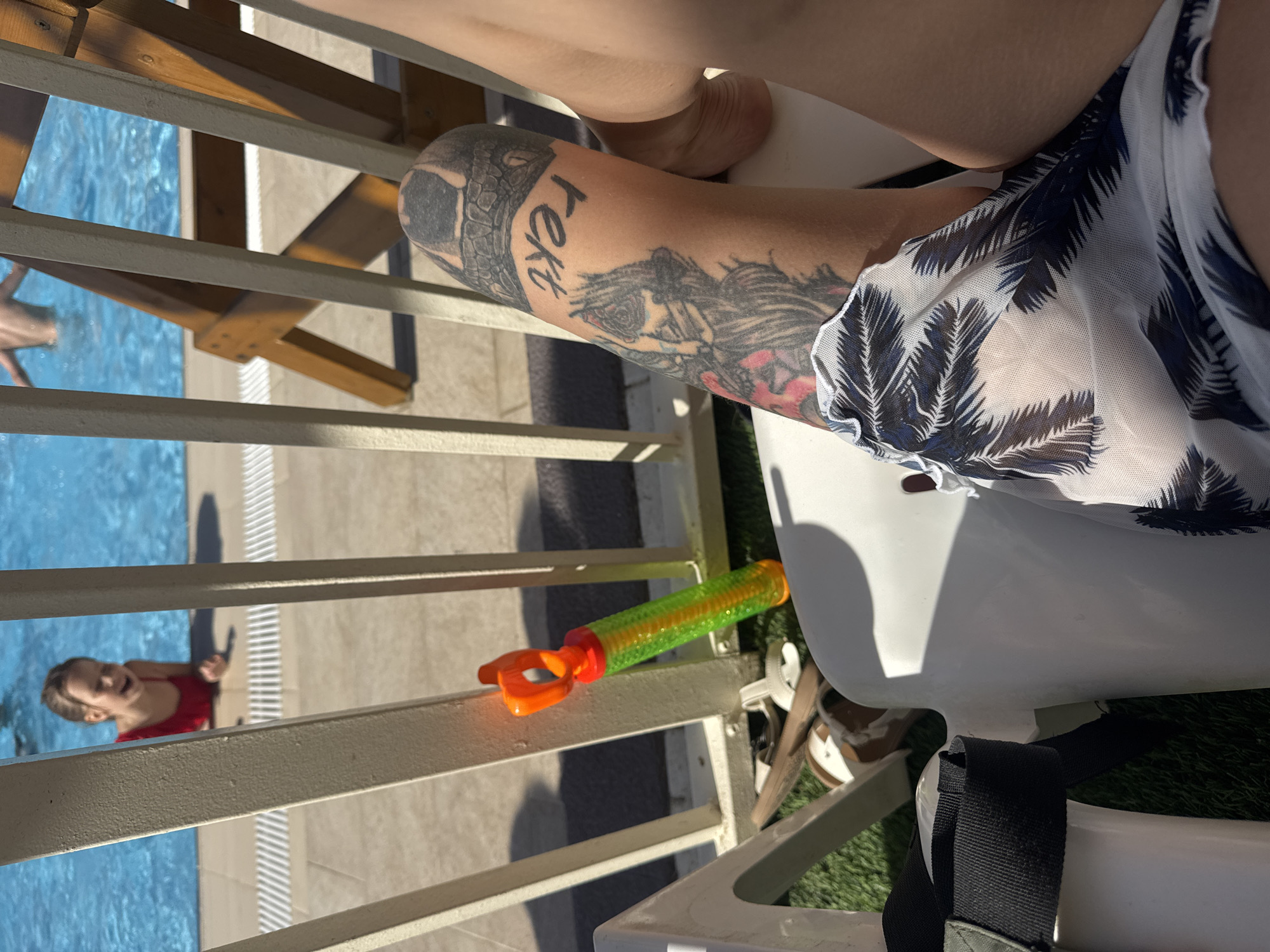The image shows a partial view of a person sitting on a white chair next to a pool. The person has a tattoo on their arm. A child in a red bathing suit is visible in the pool. A colorful plastic water gun sits on the chair next to the person. The setting is outdoors, likely near a swimming pool in a resort or recreational area. The location is indicated as Bat Yam, Israel.
