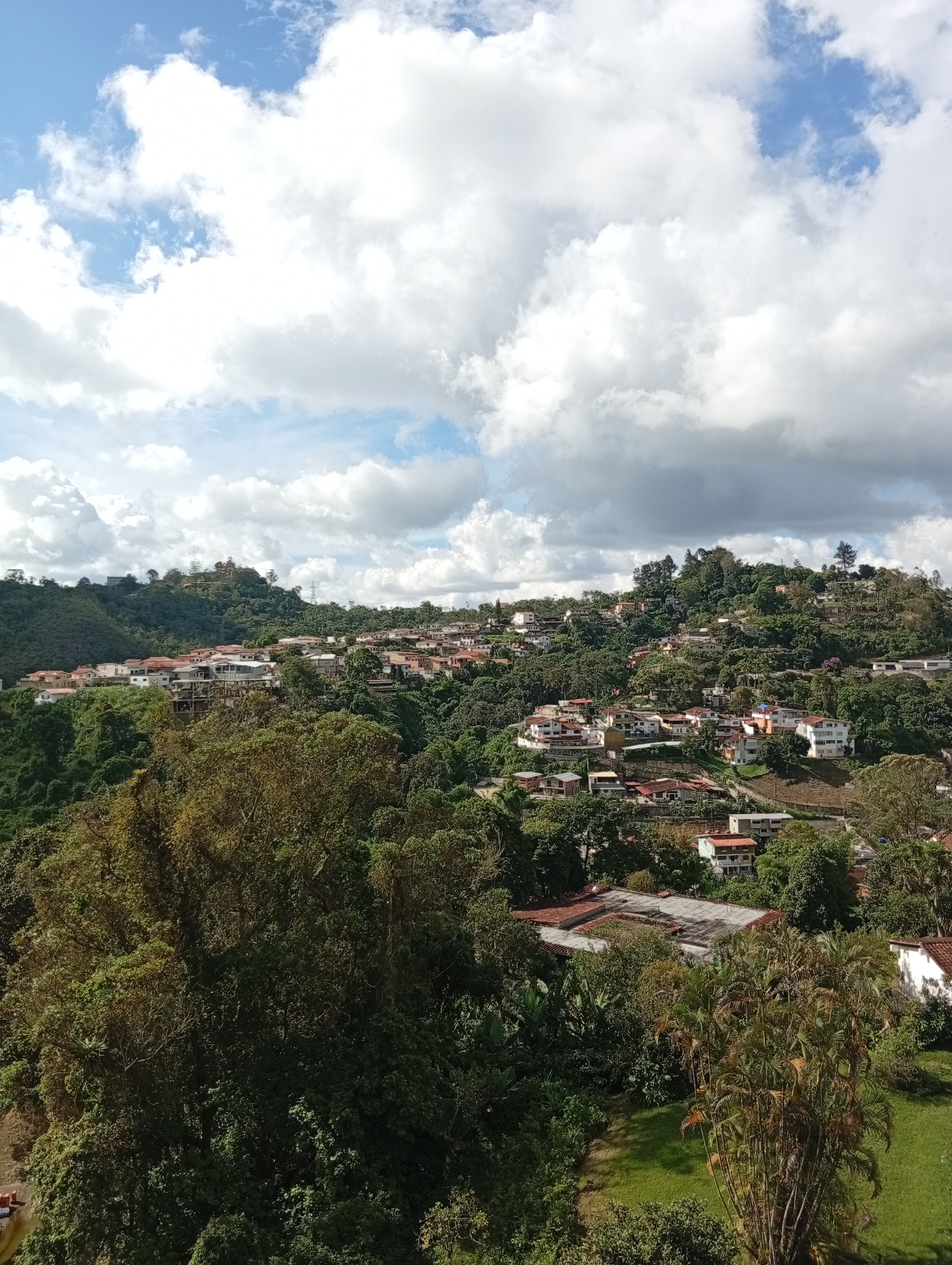 The image depicts a view of a residential area nestled on a hillside in San Antonio de Los Altos, Venezuela. The architecture of the houses features predominantly red-tiled roofs and white or light-colored walls, typical of tropical regions. The houses are spread across the undulating terrain, with dense green foliage of trees and shrubs interspersed throughout the community. The natural landscape is a prominent feature, with lush vegetation covering the hills. In the distance, on a higher elevation, a few more structures are visible, along with what appears to be a utility pole. The sky above is a mix of blue with dramatic, large, white cumulus clouds, suggesting a partly cloudy day. The overall impression is of a developed area integrated into a verdant natural environment. There are no people visible in the frame.