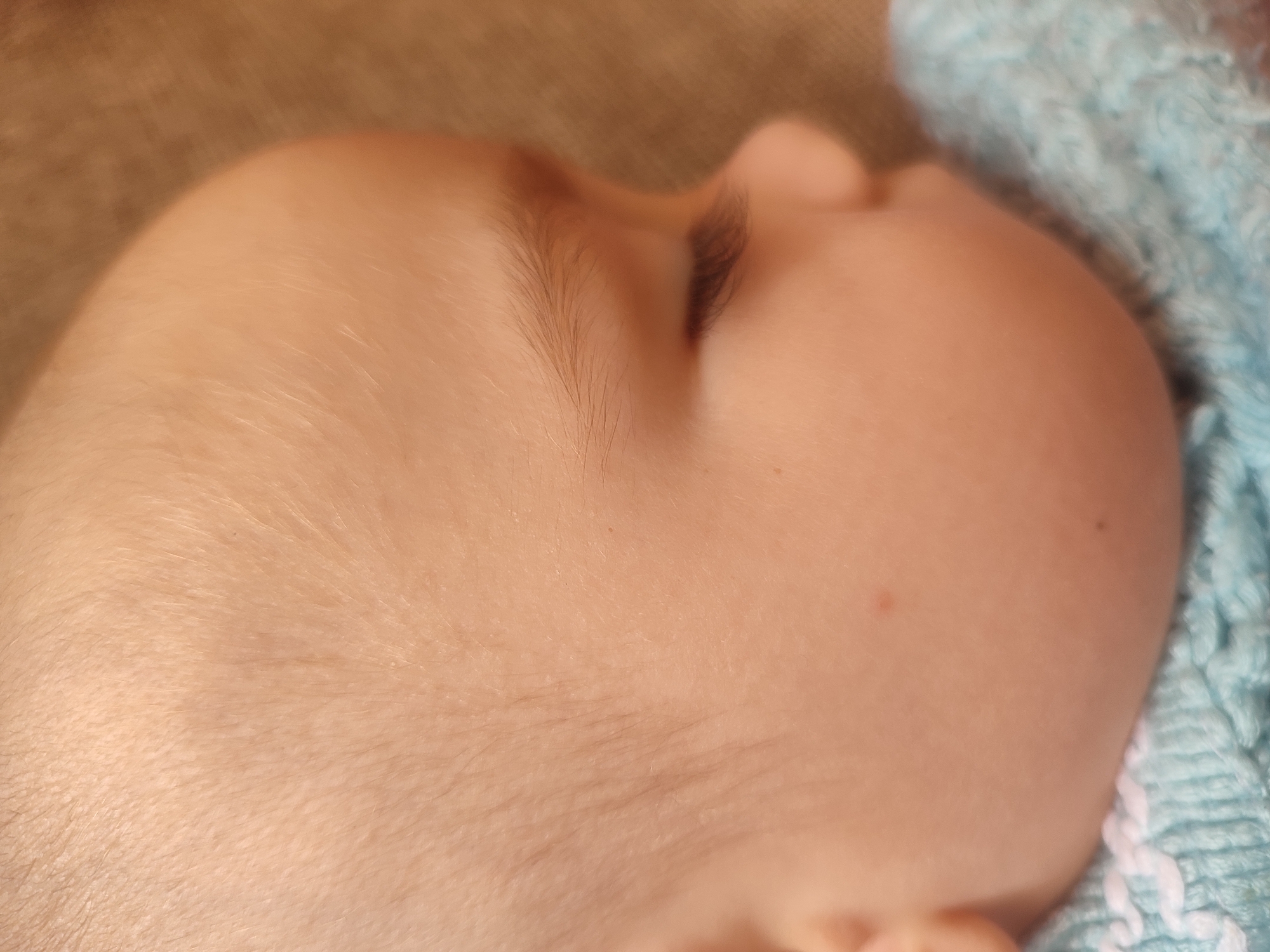 The image is a close-up, overhead shot of a baby's head, likely in Sverdlovskiy, Russia. The baby appears to be asleep, with its head turned to the right. The skin is fair and smooth, with a few wisps of fine hair visible on the scalp and a delicate eyebrow. To the right, a portion of a blue knitted blanket is visible, suggesting the baby is being held or is resting on the soft material. The background is a blurred brown texture, possibly a piece of furniture or fabric.