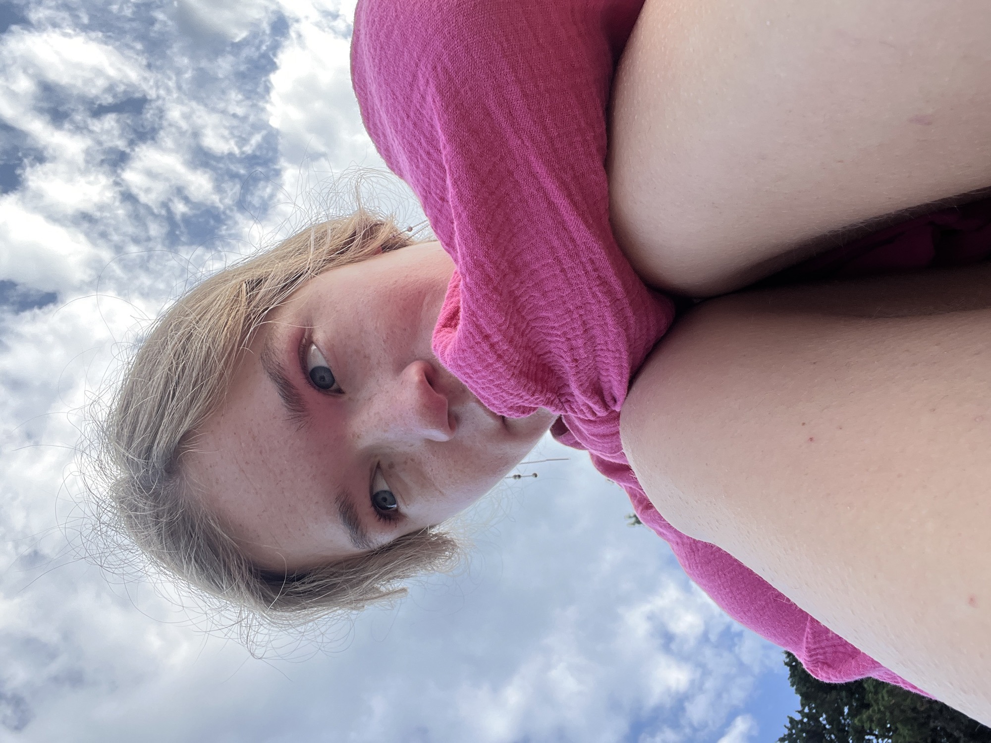 A young person with blonde hair and blue eyes looks directly at the camera from a low angle. They are wearing a pink garment that appears to be wrapped around them, possibly a shirt or blanket. The person's face is slightly flushed, and they have freckles. Behind them, the sky is visible with a mix of blue sky and white clouds. In the far bottom right corner, there is a glimpse of green foliage, possibly a tree. The photo's orientation is rotated, making the subject appear sideways. The location is unknown.
