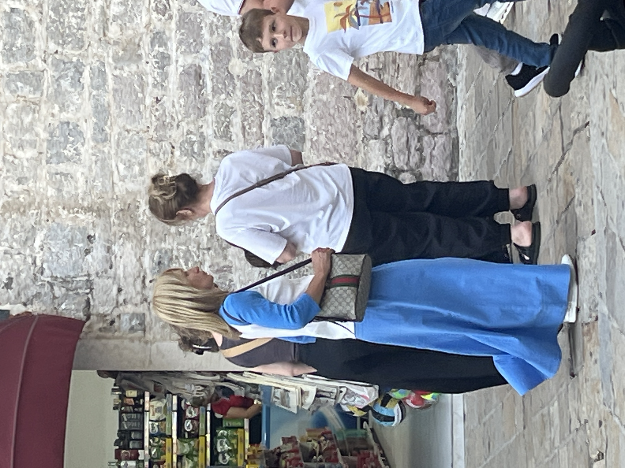 In Budva, Montenegro, a blonde woman in a white shirt with blue sleeves and a long blue skirt, carrying a Gucci crossbody bag, stands in front of a stone wall. To her left, a woman in a dark outfit is partially visible, and further left, shelves stocked with goods are seen inside a store. To the right of the blonde woman, another woman in a white t-shirt and dark pants stands facing away from the camera. A young boy in a white t-shirt with a tropical graphic and blue jeans walks past them. The scene takes place outdoors on a paved walkway, with a dark object, possibly a camera tripod, in the foreground.