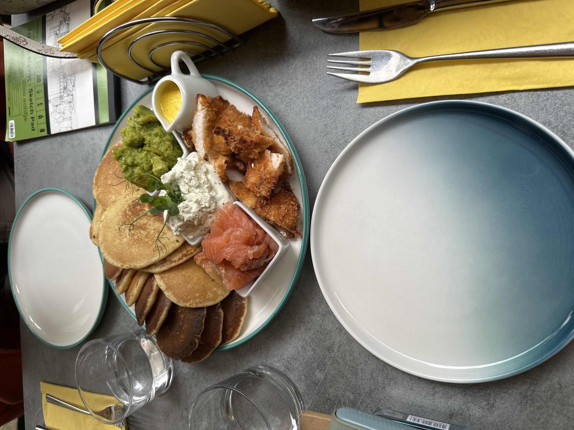 The image shows a table setting with food in a restaurant. A plate with pancakes, salmon, mashed peas, and fried chicken is in the center, along with a small pitcher of sauce and a bowl of creamy dip. Two glasses of water, a yellow napkin, a fork, and a knife are also visible. In the background, there are two white plates, a notebook, and a napkin holder. The overall scene suggests a meal is being served in a restaurant environment. The location is not determinable from the image.