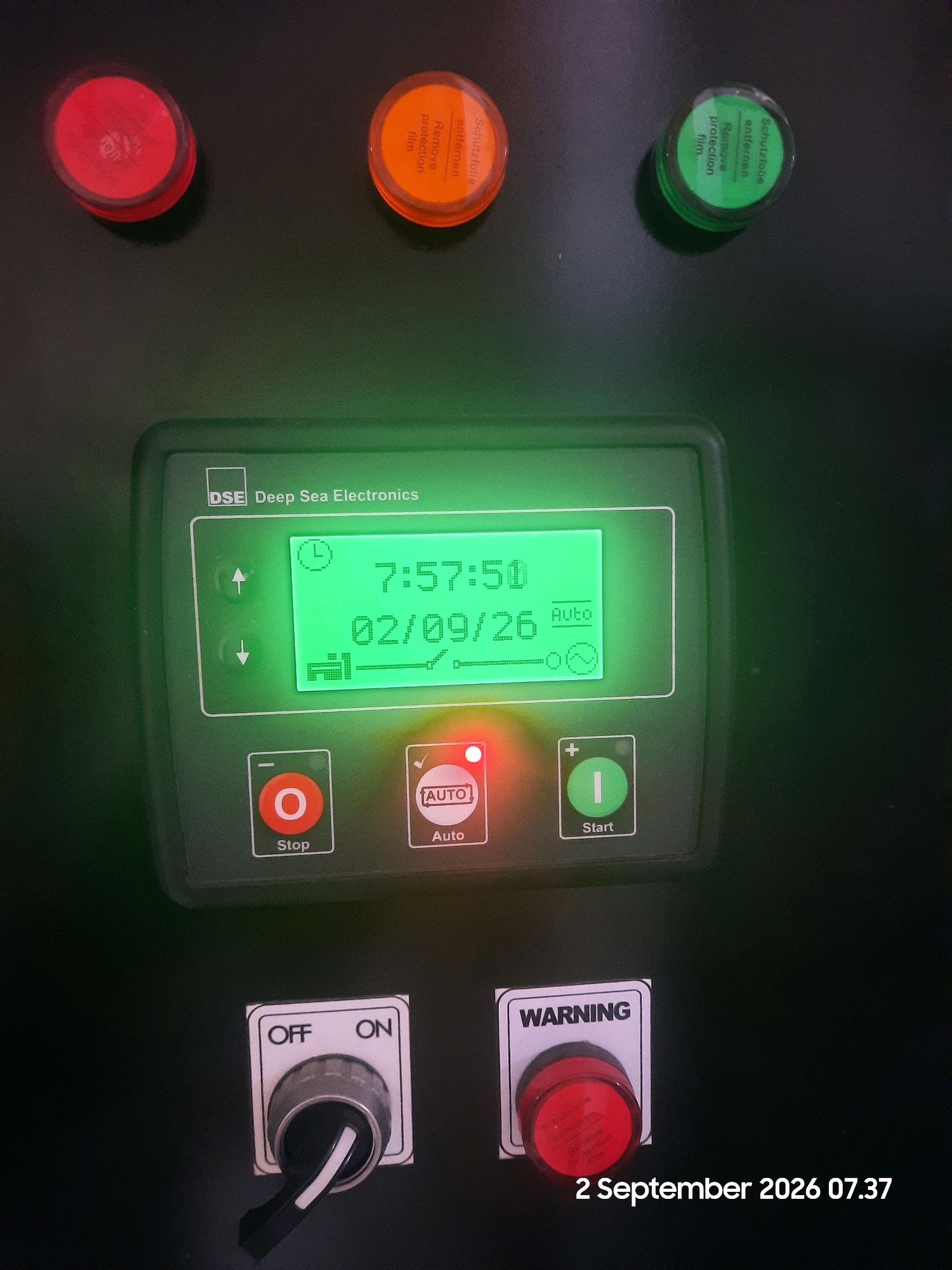 The image shows a control panel for what appears to be a generator, with various buttons, indicators, and a digital display. The control panel is mounted on a dark surface, possibly a machine or a wall.
At the top of the panel, there are three circular buttons: a red one on the left, an orange one in the middle, and a green one on the right. Each of these buttons has protective film on them with text in German and English, reading "Schutzfolie entfernen" and "Remove protection film".
Below these buttons is a digital display screen that shows "7:57" and "02/09/26" along with some icons. To the left of the display are two illuminated up and down arrows. Below the display are two larger buttons: a red "O Stop" button and a green "I Start" button with a red "AUTO" indicator illuminated.
Further down and to the left, there is a toggle switch labeled "OFF" and "ON", with the switch currently in the "OFF" position. To the right of the toggle switch, there is a red "WARNING" button.
The entire scene is illuminated by the green glow of the display and the red indicator light, giving it a somewhat artificial and technical ambiance. The image was captured on September 2, 2026, at 07:37. The image does not contain any information about the city of Gending, Indonesia, or any people present.