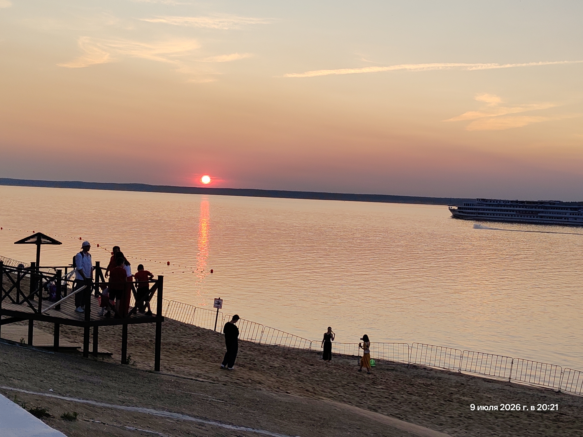 This is a photograph of a sunset over a body of water. The sun is a bright orange orb low on the horizon, casting a reflection on the water's surface. The sky is a gradient of orange, pink, and yellow. On the left side of the image, a wooden boardwalk extends out over the sand. Several people are standing on the boardwalk, some appearing to be engaged in conversation. One person is wearing a white shirt and a cap, and holding a long stick. To the right of the boardwalk, a sandy beach slopes down towards the water. A metal fence runs along the edge of the beach. Further out in the water, a large white ship is visible, steaming across the scene. On the beach, in the middle ground, three individuals are standing near the fence. One is facing away from the camera, another is looking at the water, and the third is holding a camera. The foreground shows a sandy embankment with some rough, light-colored texture, possibly a pathway or a foundation. A watermark in the bottom right corner reads "9 июля 2026 г. в 20:21", which translates to "July 9, 2026 at 20:21". The overall scene depicts a peaceful evening at a waterfront location.
