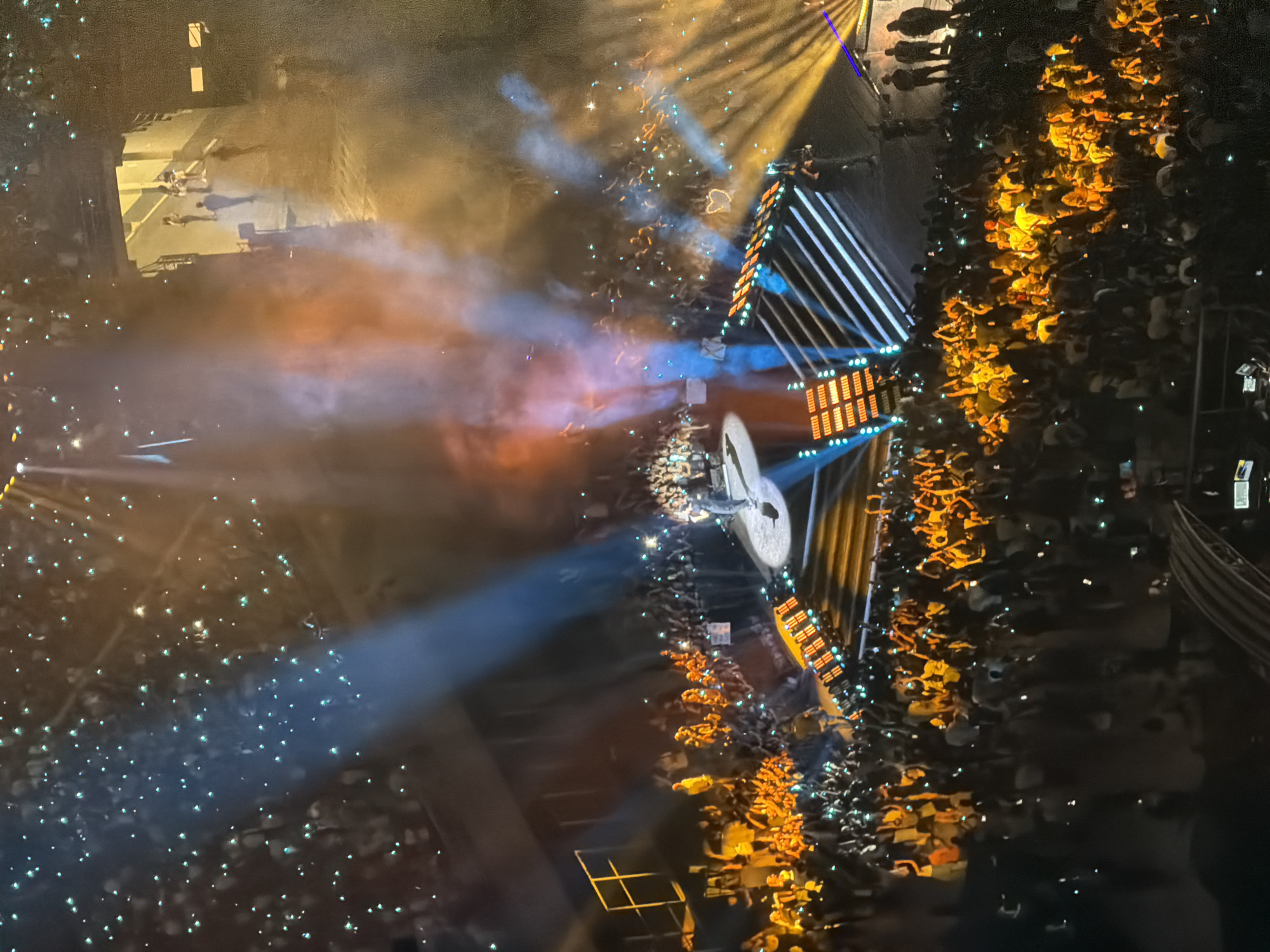 The image depicts a large, indoor concert venue in Milan, Italy, filled with an audience. A performer is visible on stage, illuminated by spotlights and surrounded by smoke effects. The crowd is engaged, with many holding up their mobile phones, their lights creating a bokeh effect within the scene. The stage itself features a dynamic lighting setup with illuminated risers and a backdrop. Sections of the audience are visible in tiered seating, and some individuals are also standing closer to the stage. The overall atmosphere is one of a lively and energetic musical performance.