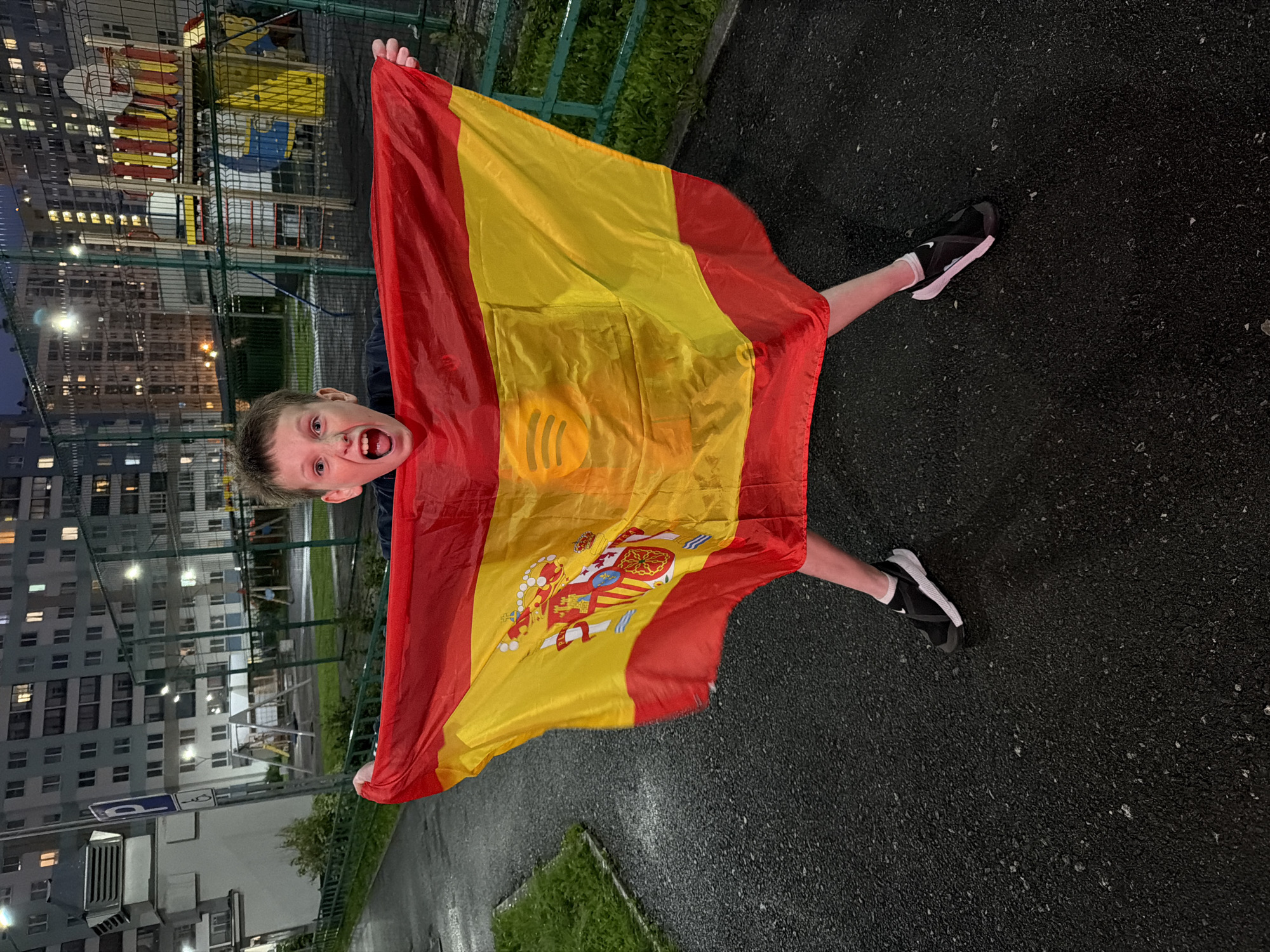 A young Caucasian male, appearing to be around 8-10 years old, stands in the foreground of an image, holding a Spanish flag. The boy's mouth is wide open in an excited expression, and his eyes are looking directly at the camera. He is wearing a dark shirt, black athletic shoes with white soles, and white socks. The Spanish flag, predominantly red and yellow, is spread out in front of him, covering his lower body and extending outwards. The flag displays the coat of arms of Spain. In the background, to the left, is a multi-story apartment building with many illuminated windows, suggesting it is evening or night. A tall, grey utility box with a white 'P' sign and a wheelchair accessibility symbol is also visible near the building. To the right of the boy and behind him is a fenced-in playground area with colorful structures, including slides and a basketball hoop. The ground in the foreground is paved with asphalt, which appears wet, possibly from rain. The overall environment suggests an urban residential area in Yekaterinburg, Russia, likely a courtyard or public space adjacent to residential buildings and a playground.