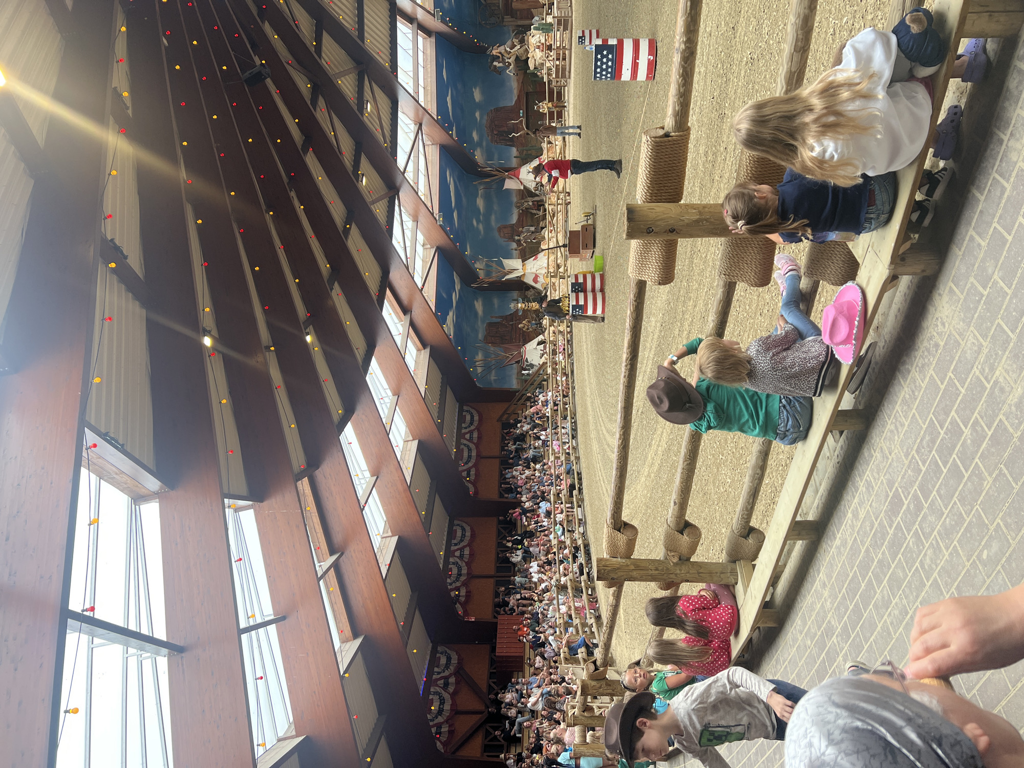 The image depicts an indoor arena with a sandy floor, likely used for equestrian events or shows, located in the city of Collendoorn, Netherlands. A large crowd of spectators, consisting of adults and children, is seated on tiered wooden bleachers on the left side of the frame. The arena is decorated with American-themed elements, including a mural depicting a Western landscape with tepees and cowboys, as well as barrels painted with American flags. In the center of the arena, two performers are visible, one in a red shirt and black pants, and another in a dark shirt and jeans, appearing to be engaging the audience. Several children are seated on a bench in the foreground, facing the arena and watching the show. The arena has a sloped, wooden ceiling with skylights and is adorned with strings of red and yellow lights. The overall atmosphere suggests a lively entertainment event.