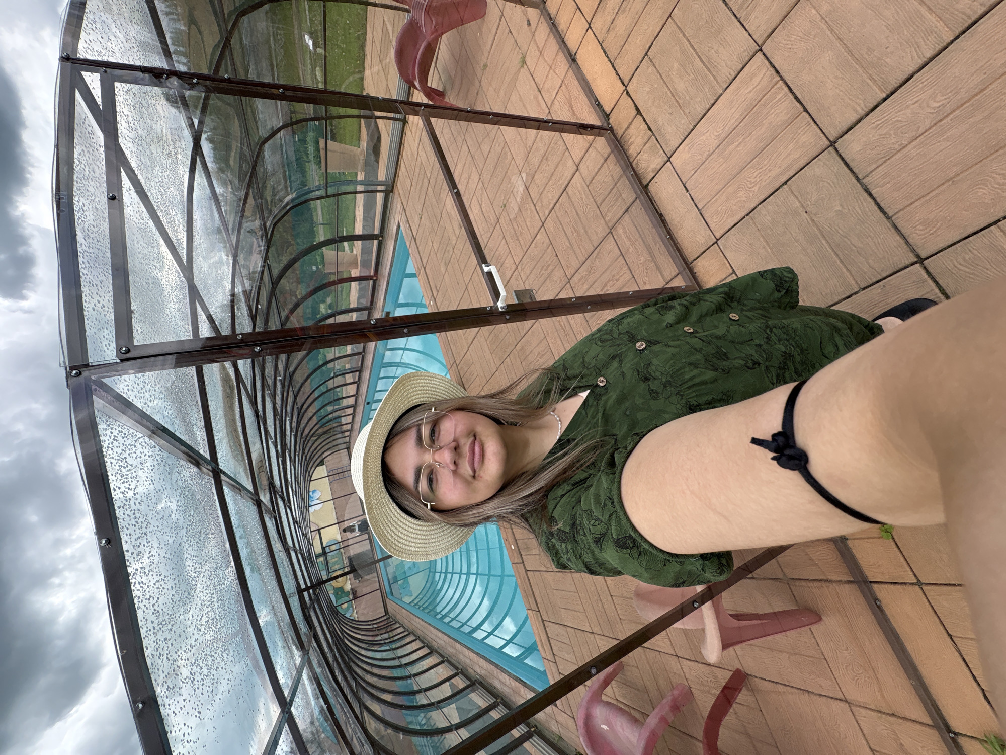 A young woman with long, light brown hair, wearing a straw hat, glasses, and a green patterned button-up shirt, poses for a selfie indoors. She is standing on a wooden tiled floor next to a covered swimming pool. The pool is enclosed by a transparent, arched structure with a metal frame. The sky outside appears cloudy. The woman has a black band tied around her right arm. In the background, there are plastic chairs and potted plants. The environment suggests a recreational facility, possibly a hotel or a public swimming area. The location is stated to be Birobidzhan, Russia.