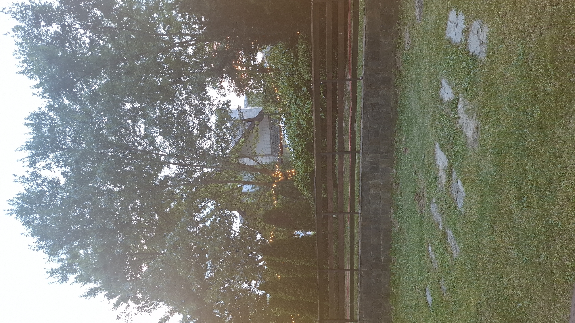This is a photograph taken in Razlog, Bulgaria. The scene appears to be an outdoor area, possibly a backyard or garden, at dusk or early morning. A large tree with dense foliage dominates the left side of the frame, its branches reaching upwards and partially obscuring the sky. In the background, a building with a white facade and a grey roof is visible, surrounded by greenery. A string of fairy lights is draped across the trees and bushes, adding a warm, inviting glow to the scene.
A wooden fence with dark vertical planks separates the grassy area from the trees and building. To the right of the fence, a path made of irregularly shaped stepping stones is laid out on a green lawn. The stones are a light grey color and are scattered across the grass, suggesting a naturalistic design. The overall impression is one of a peaceful and serene setting, possibly prepared for an evening gathering or celebration. The lighting suggests a low-light condition, with the fairy lights providing artificial illumination against the natural twilight. No people are visible in the image. No text is discernible.