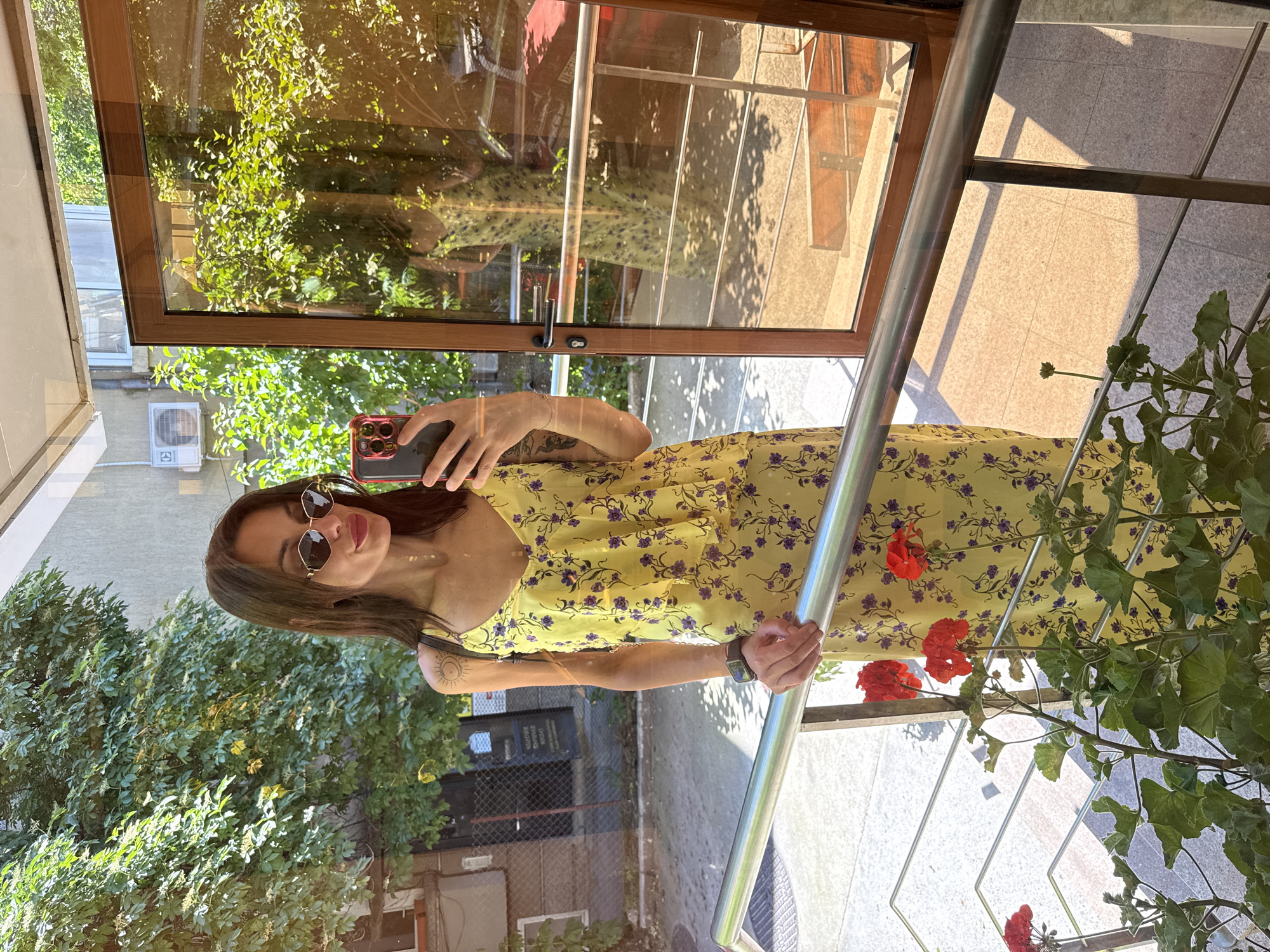 A woman is taking a selfie in what appears to be a doorway or entrance. She is wearing a yellow floral dress and sunglasses, holding a smartphone in her right hand. Her left arm is visible, showing a tattoo near her shoulder. The background includes foliage, suggesting an outdoor or garden setting. The doorway has a wooden frame and a metallic railing. The reflection in the glass shows elements of the surroundings, including building facades and greenery. The location is Pleven, Bulgaria.