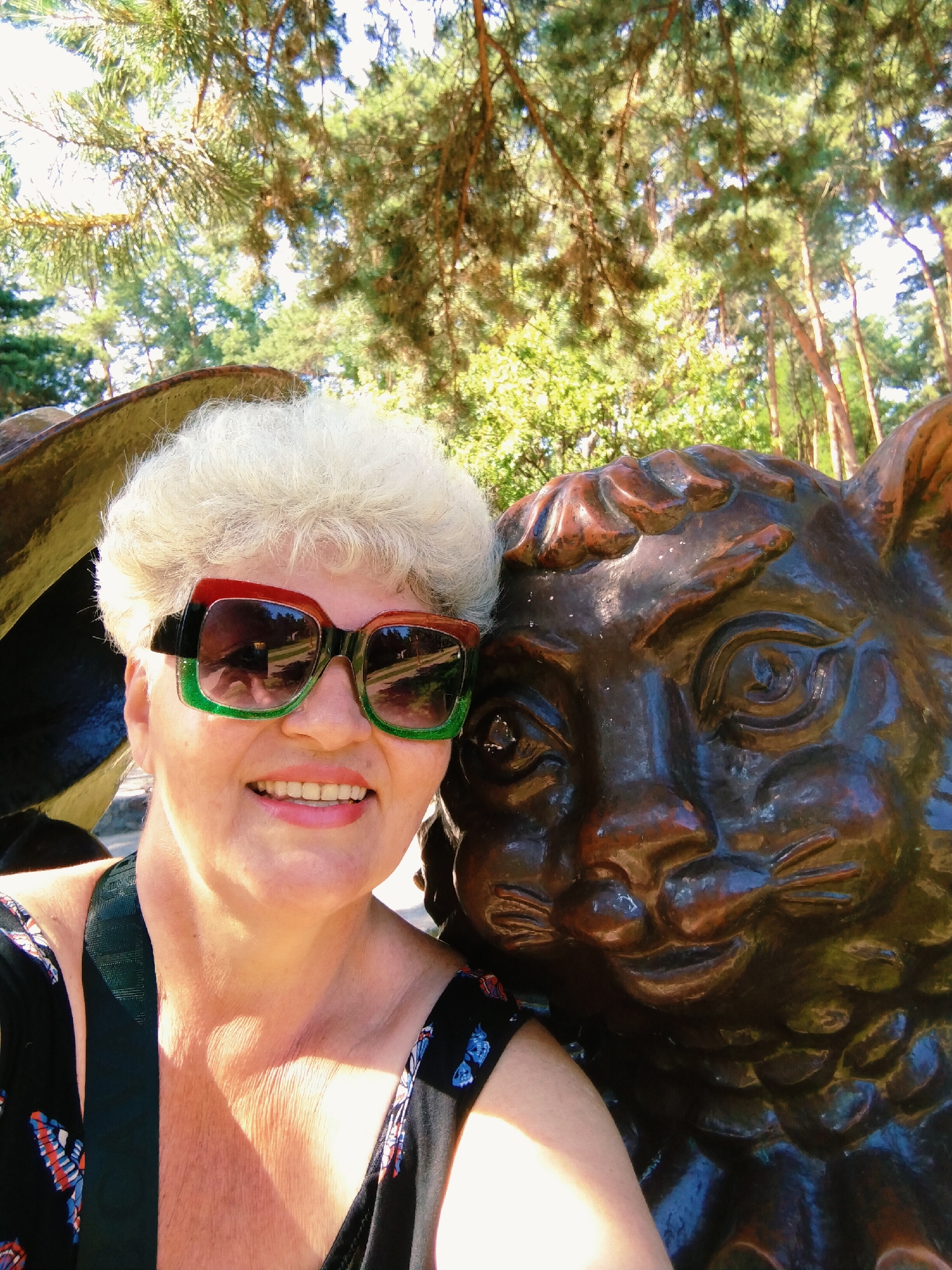 A woman with white hair and sunglasses is taking a selfie outdoors with a bronze statue of a lion. The woman is smiling and wearing a black dress with a butterfly print. She has on large, square-shaped sunglasses with red, green, and black frames. The lion statue is dark brown and appears to be part of a larger sculpture. They are positioned in front of a backdrop of tall trees, suggesting an outdoor setting in a park or forested area. The lighting indicates it is daytime, with sunlight filtering through the leaves of the trees. The scene is likely in Horishni Plavni, Ukraine, a city known for its industrial landscape and some green spaces.