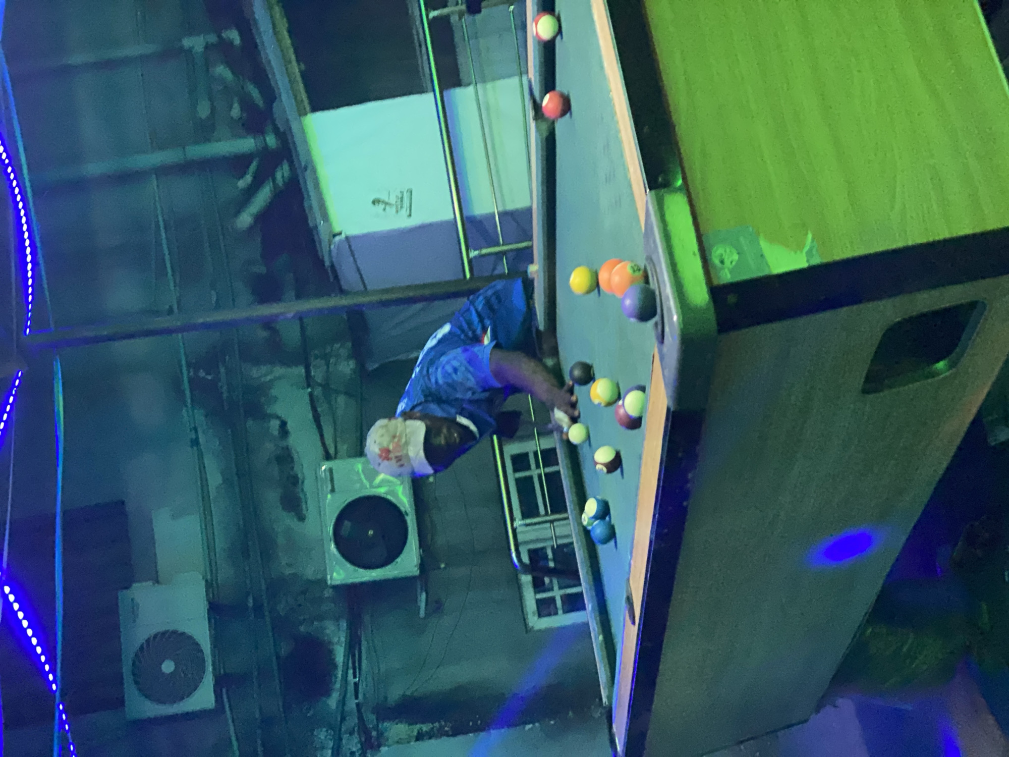 The image depicts a man playing pool inside a building, likely in a recreational establishment. The location is Ado, Nigeria.
**People:**
One adult male is visible, wearing a blue patterned shirt and a white head covering, appearing to be lining up a shot on a pool table.
**Subjects:**
The primary subject is the man playing pool.
**Objects:**
A pool table with multiple billiard balls, a cue stick (implied, not visible), an air conditioning unit mounted on the wall, a window with multiple panes, blue LED light strings and a white sign with a small silhouette on it.
**Actions:**
The man is in the act of playing pool, focusing on his next shot.
**Interactions:**
The man interacts with the pool table and billiards, aiming to strike the cue ball.
**Environment:**
The scene appears to be set indoors, possibly in a game room or a recreational space. The lighting appears to be a combination of fluorescent and LED lighting, as indicated by the hue of the room. The room shows signs of being a commercial space.
