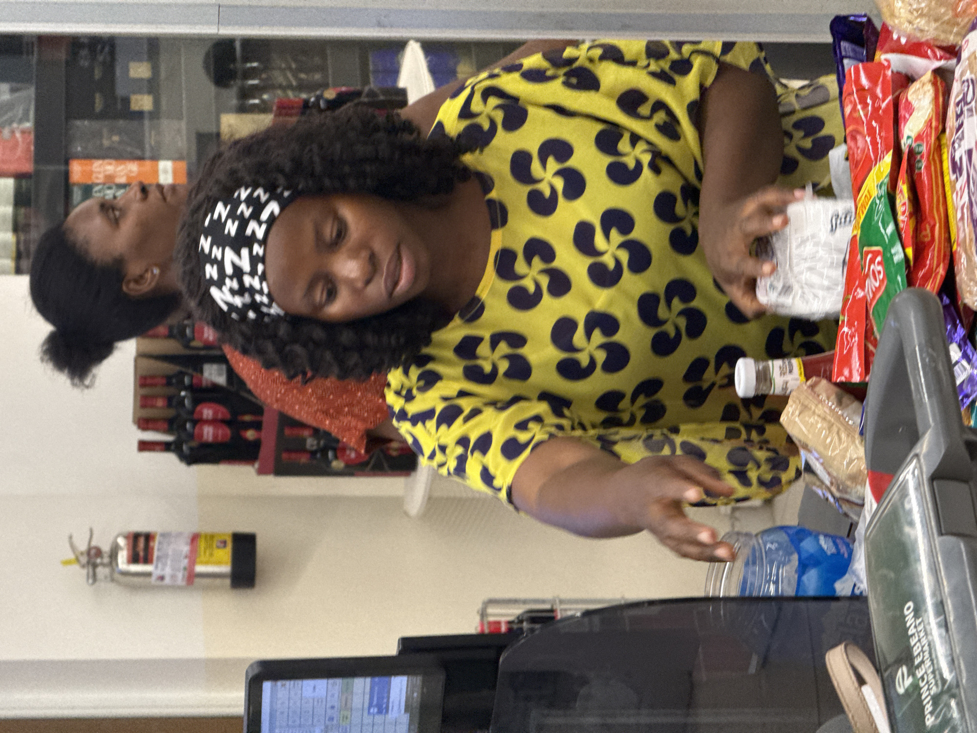 Two women are at a checkout in Prince Ebeano Supermarket. The woman in the foreground has dark curly hair, a black and white headband, and is wearing a yellow top with dark blue swirling designs. She is reaching for an item near the cash register. The other woman has her hair pulled back and is looking off to the side. A fire extinguisher is mounted on the wall. Various grocery items are visible on the conveyor belt and in the background, including bottles of wine. The location is likely Abuja, Nigeria.
