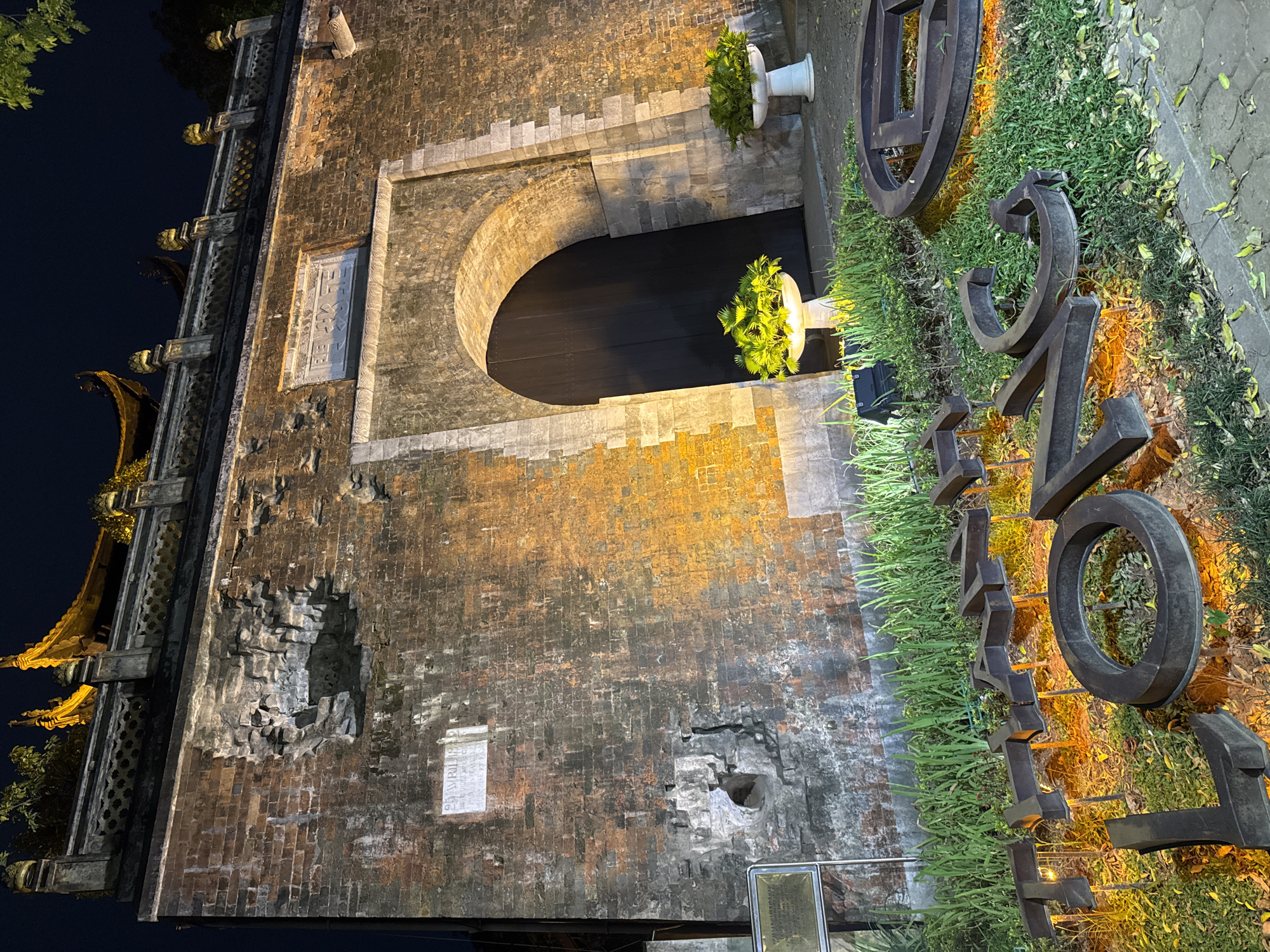 The image shows the entrance gate of the Imperial Citadel of Thang Long in Hanoi, Vietnam, at night. The ancient brick wall of the citadel, illuminated by yellow lights, features a large arched doorway with a dark wooden door. The wall has visible signs of age and damage, with sections of brick missing. Above the archway, there is a stone plaque with inscriptions. The rooftop of the structure is visible in the upper portion of the image, adorned with traditional Vietnamese architectural elements. In front of the gate, large metallic letters spelling out "THANG LONG" are placed on the ground amidst grass and fallen leaves. Two white planters with green foliage stand on either side of the entrance, and small spotlights illuminate the lettering. The scene is lit by artificial lights, creating a dramatic effect against the dark night sky.