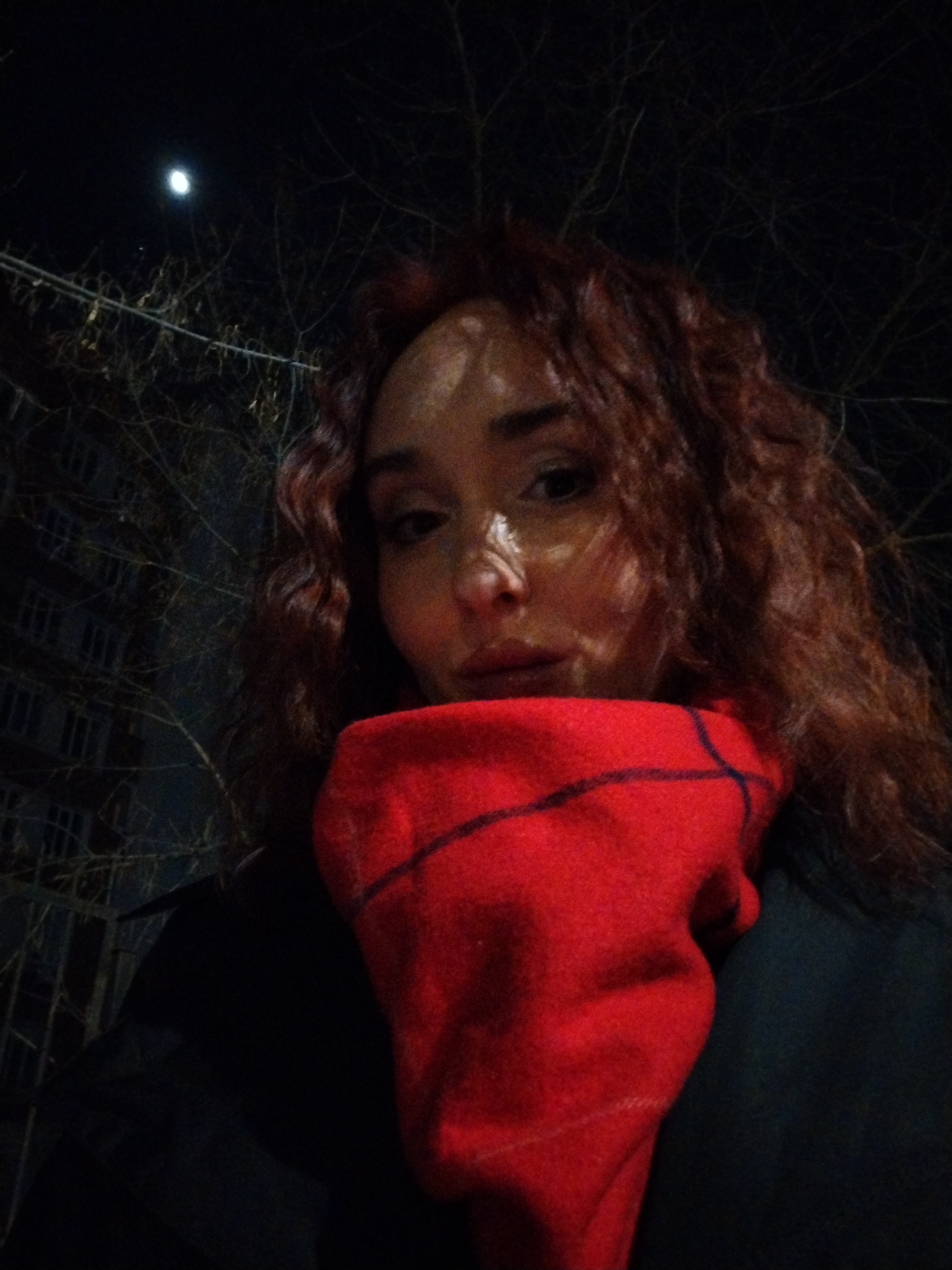 The image displays a nighttime close-up of a human subject, appearing female, positioned outdoors in Izhevsk, Russia. The subject occupies the majority of the frame, facing forward with their eyes directed towards the point of capture. They have long, wavy, reddish-brown hair and are wearing a dark outer garment along with a bright red scarf or neck warmer patterned with a dark grid. The subject's face is illuminated from the left side (viewer's left), creating highlights and shadows across their features. In the upper left background, a crescent moon is visible against a dark sky. Bare tree branches are discernible throughout the upper half of the background, and to the left of the subject, a multi-story building with visible window frames is partially silhouetted.