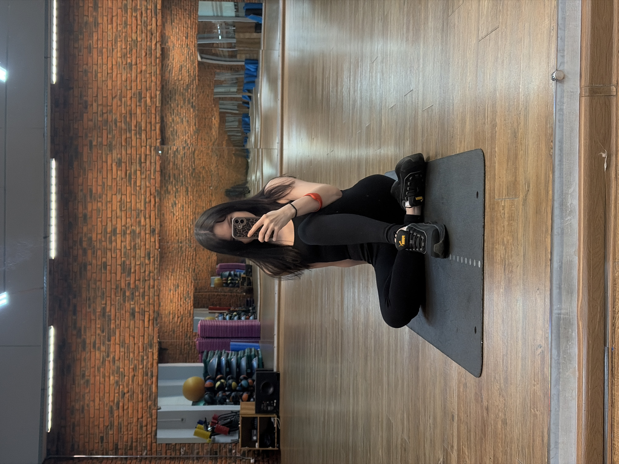A young woman wearing black athletic clothing is kneeling on a grey yoga mat in a gym. She holds a smartphone in her left hand, seemingly taking a photo in a mirror. The gym has brick walls on one side and a wooden floor. Exercise equipment, including yoga mats and weights, is visible in the background, reflected in the mirror. The location is in the city of Chaikovsky, Russia.