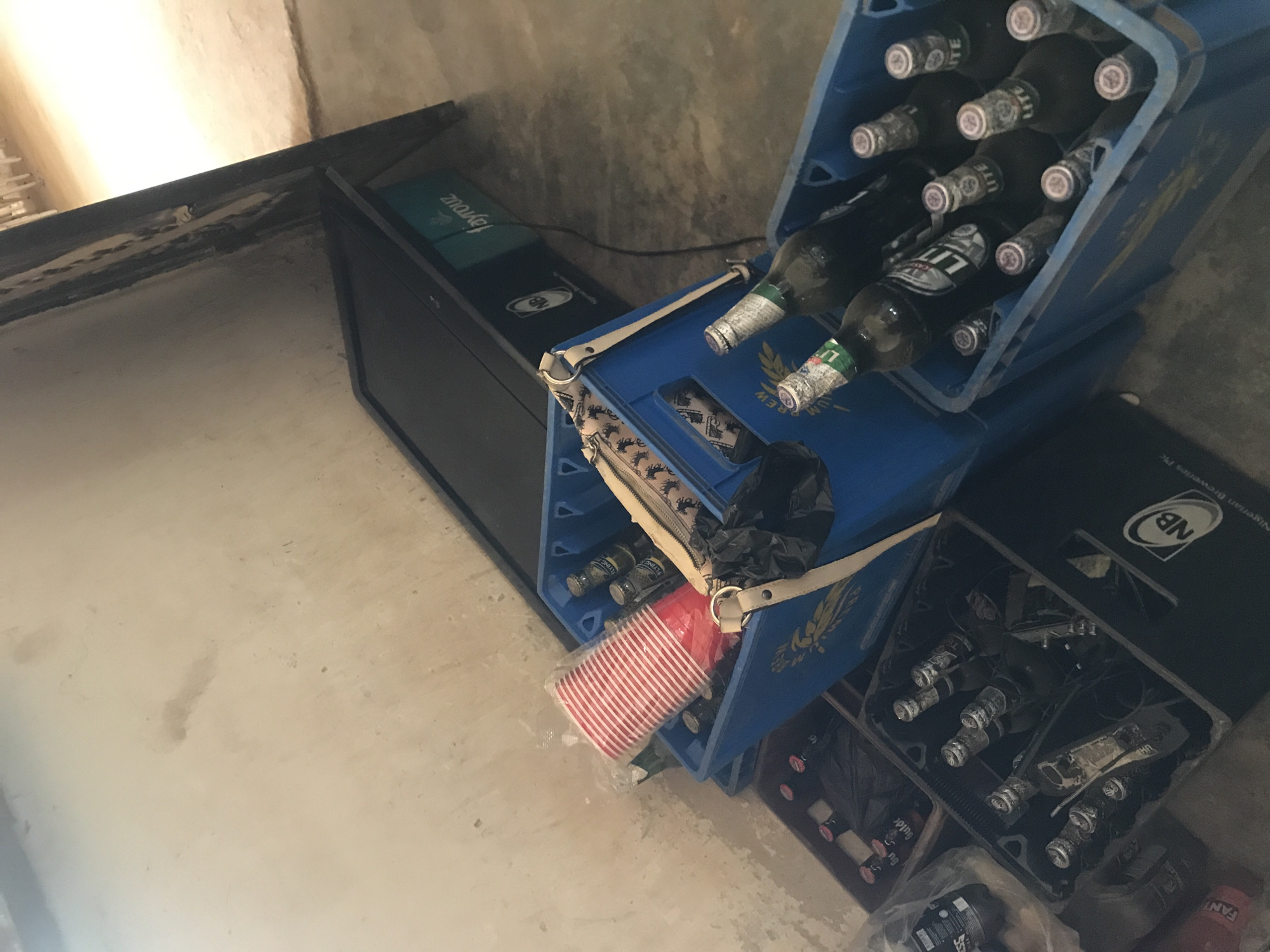 The image shows a scene of what appears to be an informal gathering or storage area for beverages, possibly in New Stadium, Nigeria. In the foreground, several blue plastic crates are filled with bottles of "LITE" beer. One crate contains at least ten bottles, with some obscured from view. Another blue crate, positioned slightly behind and to the left, holds a stack of red and white striped disposable cups within a plastic wrapper, as well as several bottles of beer. To the left of this crate, a black crate is visible, also containing beer bottles, some of which are covered by black plastic bags. Further to the left, a transparent plastic bag contains more bottles, one of which is red and labeled "FANTA". In the background, a dark, rectangular object, possibly a screen or a large electronic device, is propped against a wall. The environment appears to be an indoor or semi-enclosed space with a concrete floor and light-colored walls, suggesting a utilitarian or storage area. There are no people visible in the image.