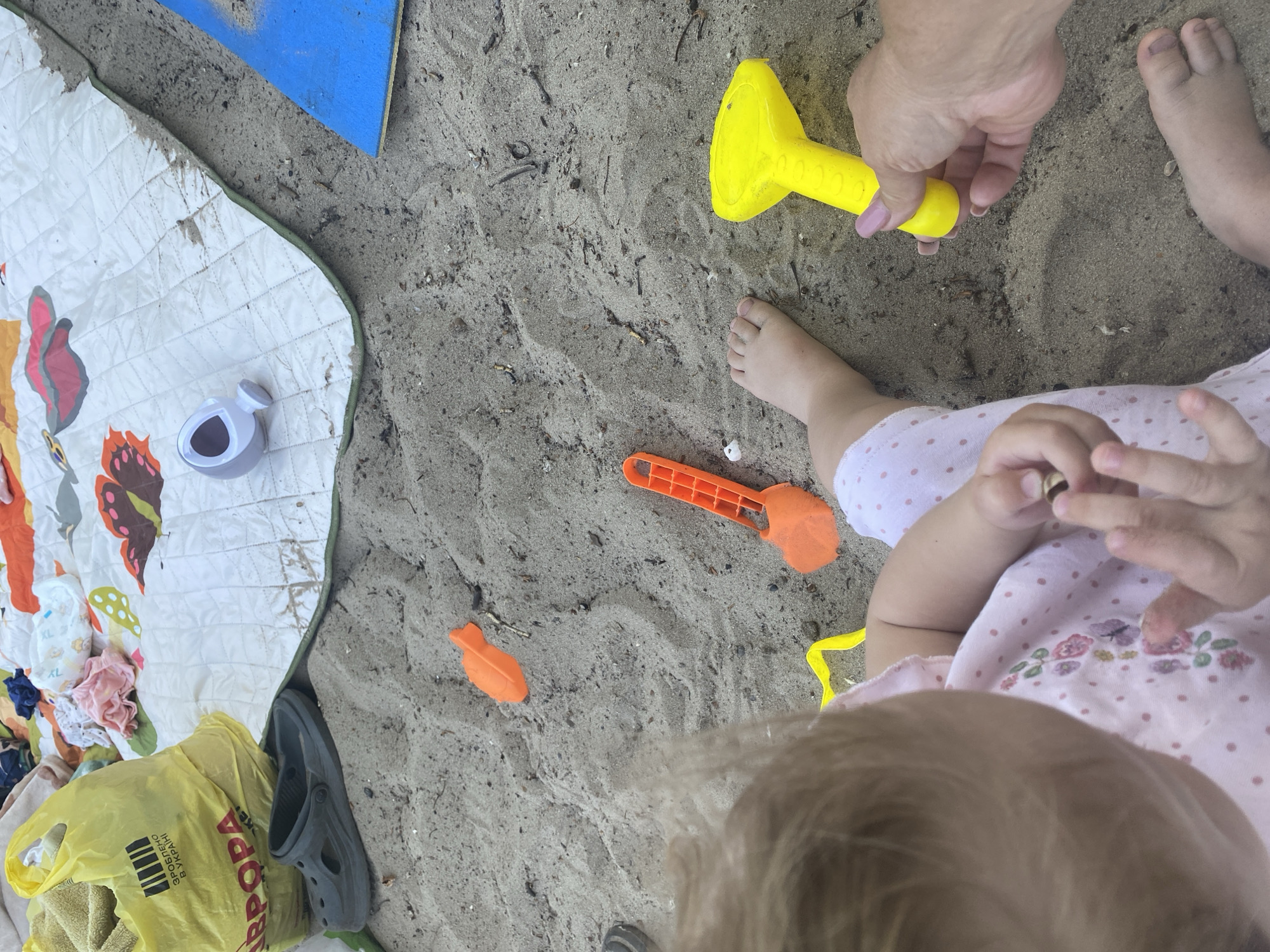 The image shows a child's feet and hands playing in sand. A yellow plastic shovel and an orange plastic shovel are visible, along with an orange toy fish. A person's hand is holding the yellow shovel. In the background, a picnic blanket with butterfly designs is spread out on the sand, and a yellow plastic bag is partially visible. A dark gray Croc shoe is also present. The scene appears to be a sandy playground or beach, possibly in the city of Dnipro, Ukraine.