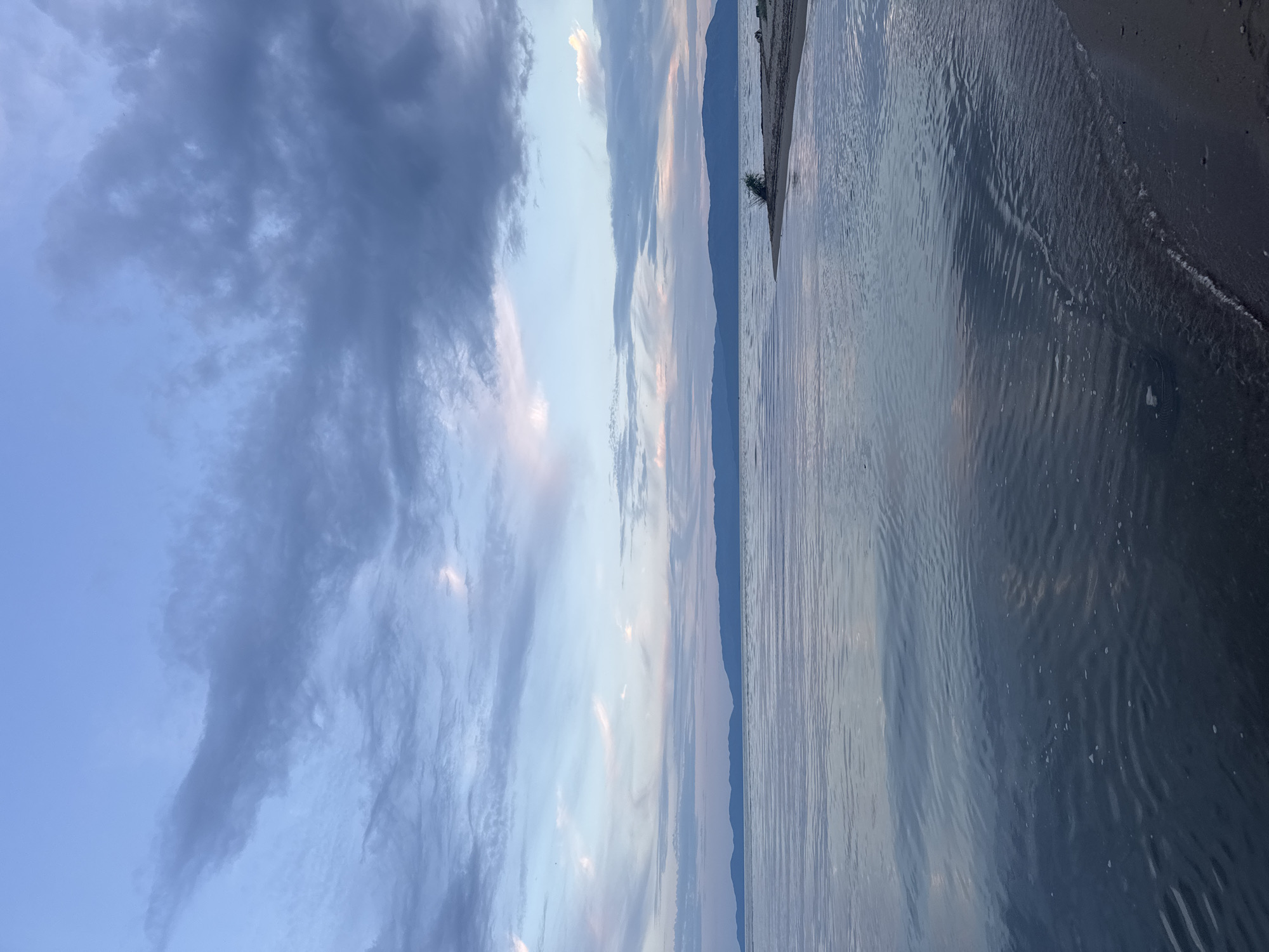 The image depicts a serene body of water at dusk, with mountains in the distance. The sky is filled with a mix of soft pink and gray clouds against a pale blue backdrop. The water's surface is calm, with gentle ripples reflecting the colors of the sky. A narrow strip of sandy beach is visible on the right side of the frame, with a small clump of vegetation. The overall scene suggests a peaceful, natural landscape.