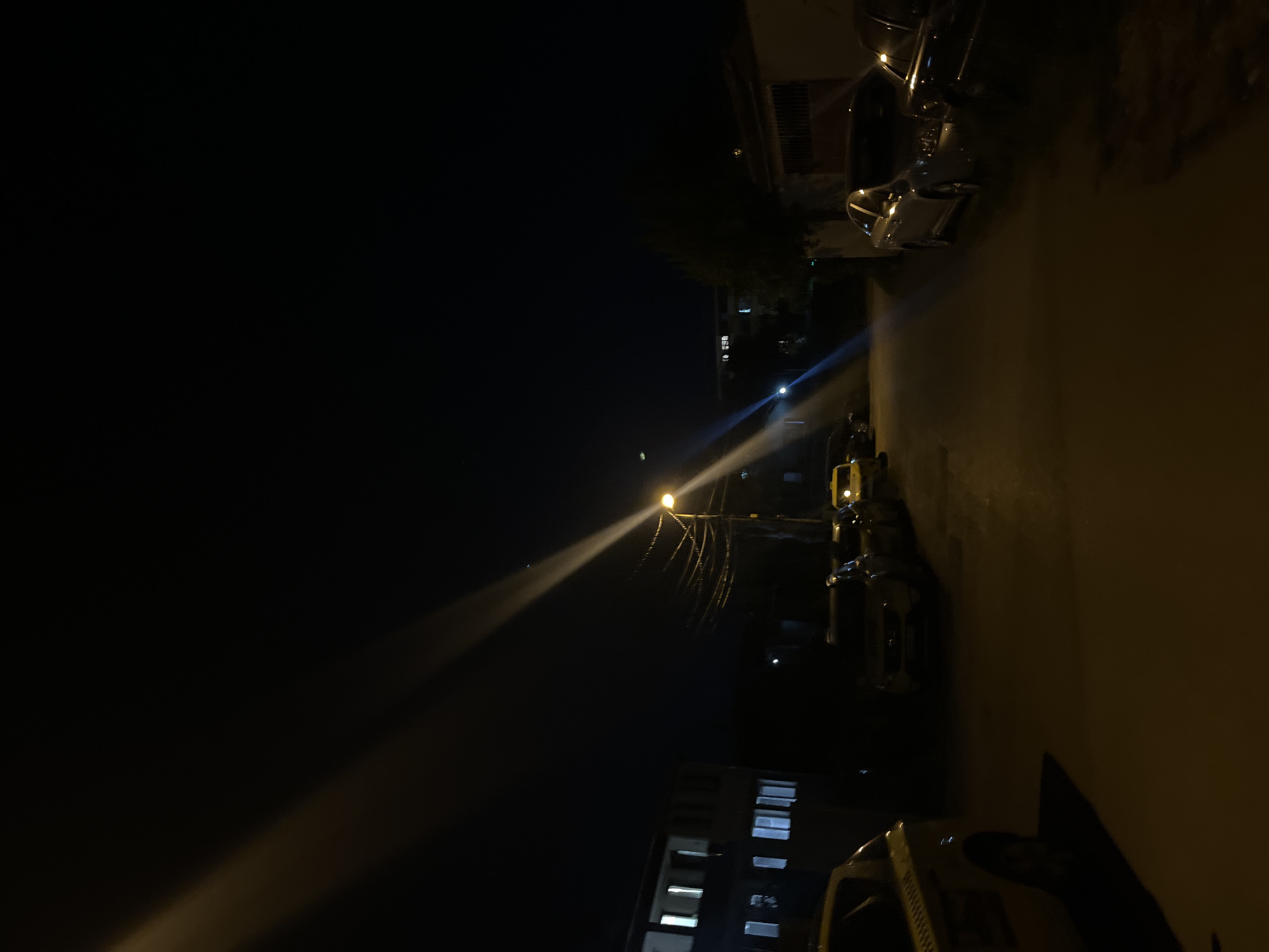 This night-time image, taken in Sofia, Bulgaria, depicts an urban street illuminated primarily by a single, prominent streetlamp. The streetlamp, positioned slightly right of the vertical center, emits a strong, yellowish-white light that forms a distinct, wide beam extending diagonally across the left side of the frame. This light pierces the deep darkness of the night, highlighting what appears to be dust or moisture in the air.
Along the right side of the image, several vehicles are visible. In the immediate foreground, part of a light-colored or yellow vehicle, possibly a taxi or commercial van, is parked. Further down the street, other vehicles, darker in color, are also present, reflecting some of the ambient light. Buildings with scattered illuminated windows can be seen on both sides of the street, receding into the background. Near the bright streetlamp, a network of utility wires or cables is visible. The overall scene is devoid of people and active events, conveying a sense of quiet solitude characteristic of a city street after dark. No discernible text is visible.
