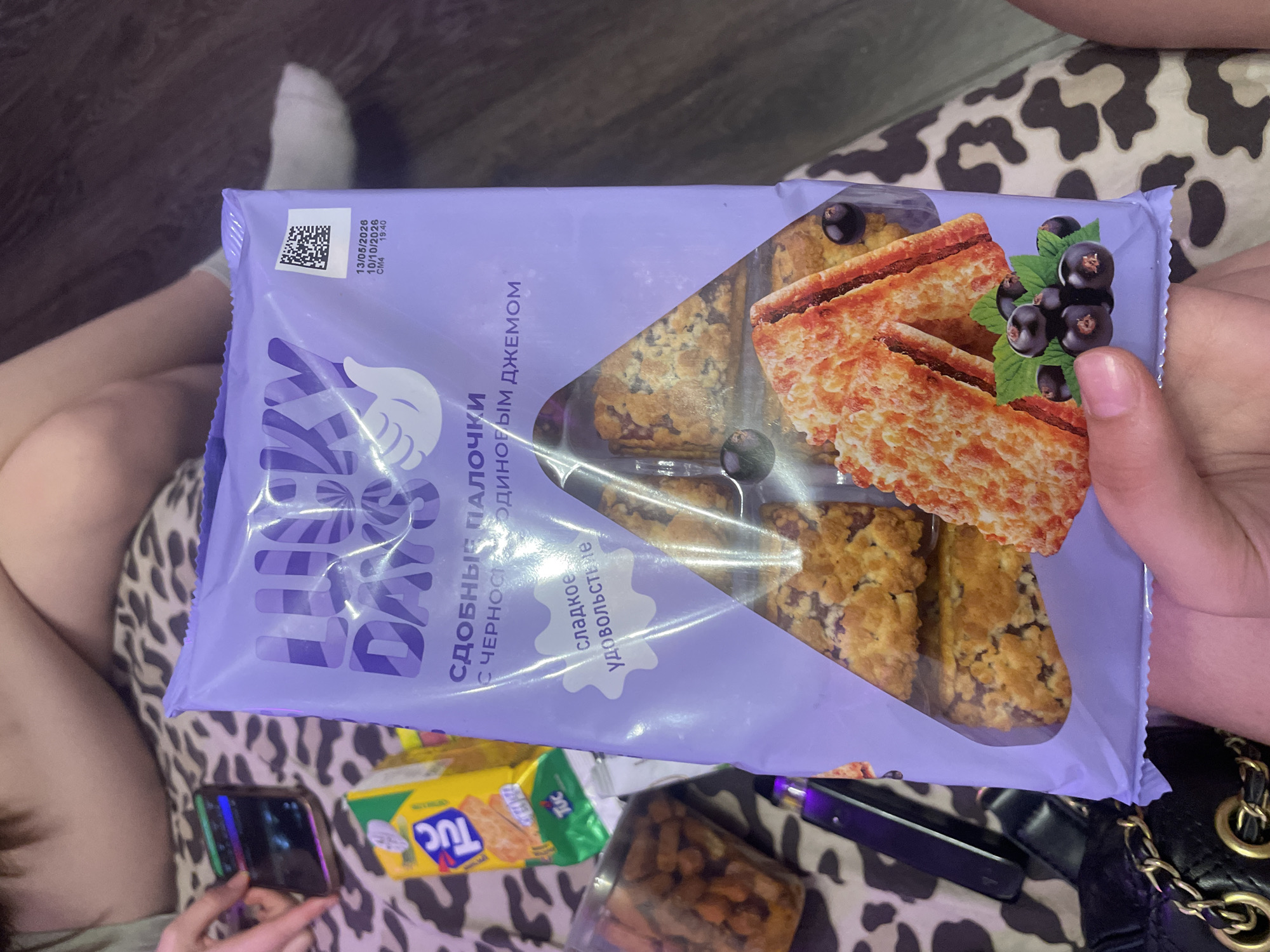 The image shows a close-up view of a purple package of "Lucky Days" cookies with blackcurrant jam. The package is held by a person's hand in the foreground. Inside the transparent window of the package, several cookies are visible, some appearing to be sandwich cookies with jam and a crumble topping, and others showing a more solid layer with jam. There is an illustration of blackcurrant berries and leaves on the lower right side of the package. To the left of the cookie package, a yellow box of "TUC" crackers is partially visible. In the background, out of focus, a person's legs and feet are visible, suggesting they are sitting or lying down. The environment appears to be indoors, possibly a casual setting like a living room or bedroom, with a patterned surface underneath. No specific location in Zmeinyi, Russia, is identifiable from the image.