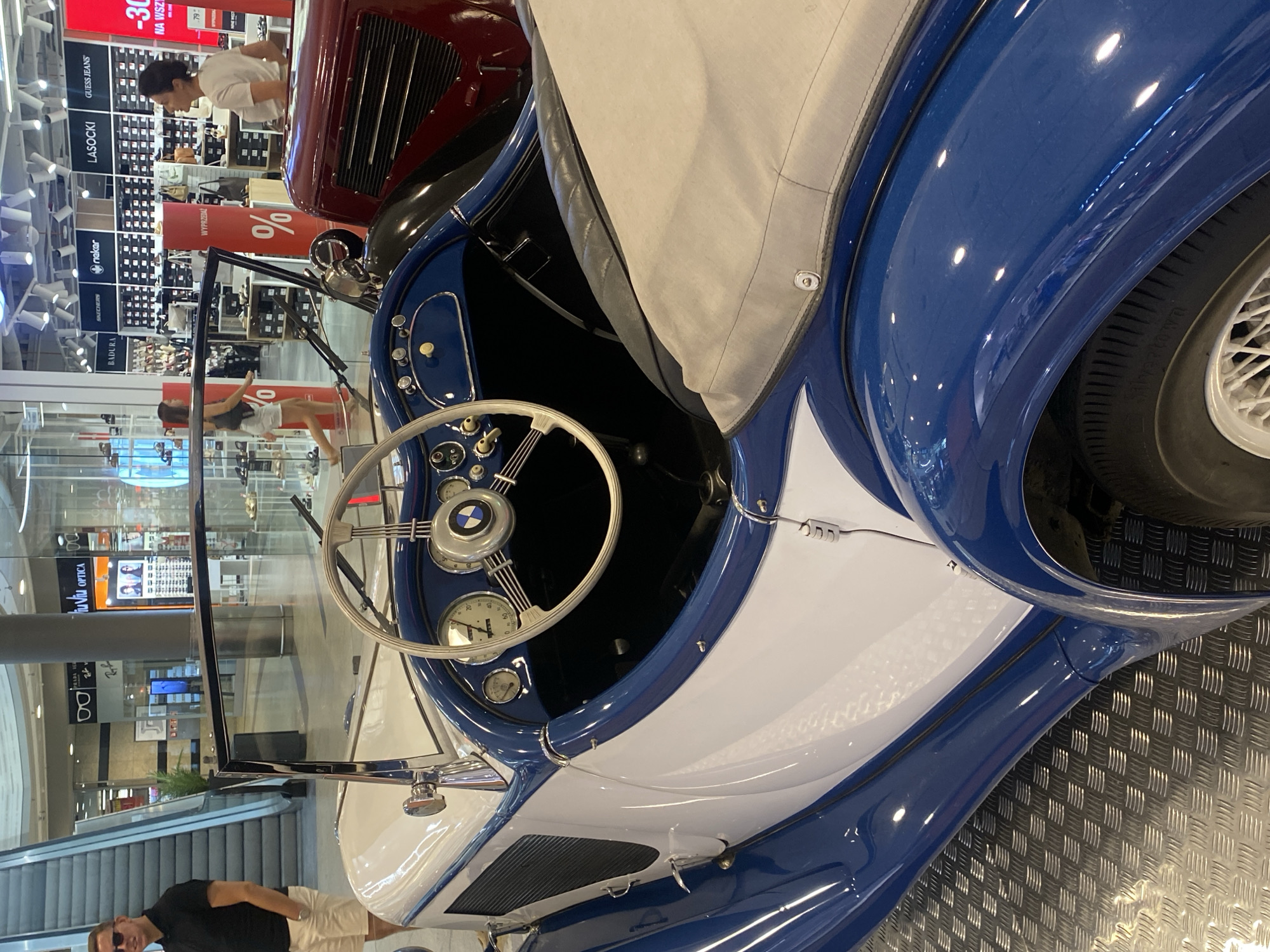 The image depicts a white and blue vintage convertible BMW car displayed indoors, possibly in a shopping mall in Łódź, Poland, given the Polish signage visible in the background ("Wypożyczalnia", "%", "na wszystko"). The car is positioned on a metallic checkered floor. In the background, retail store displays are visible, including optical shops with eyewear and clothing stores featuring brand names like "SKECHERS", "NAOKO", "LASOCKI", and "GUESS JEANS".
On the left side of the frame, a man wearing sunglasses, a black polo shirt, and light-colored shorts stands near an escalator. In the mid-ground, two women are visible. One woman, wearing a dark top and shorts, walks away from the camera. Another woman in a white t-shirt looks towards the retail displays. The car's interior, including the steering wheel, dashboard with gauges, and a portion of the beige convertible top, is prominent in the foreground. The overall scene suggests a public exhibition or a display within a commercial space.