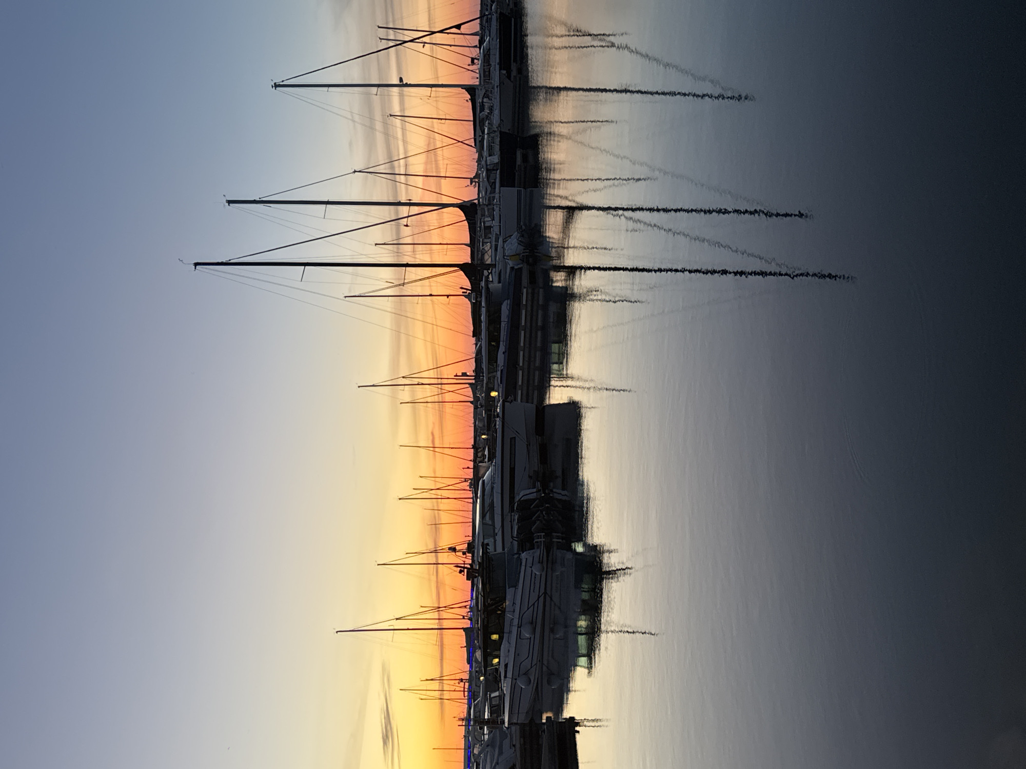 A vertical, full shot captures sailboats and motorboats docked in a marina against a twilight sky, likely in Tallinn, Estonia, given the context. The sky transitions from a pale blue at the top to vibrant orange and yellow hues near the horizon, indicating either sunrise or sunset. Numerous boats, predominantly sailboats with tall masts, are lined up and reflected in the calm, dark water. Some of the motorboats have interior lights illuminated. The overall scene is serene and peaceful, with the reflections of the boats and masts creating a symmetrical visual effect on the water's surface. There are no people visible in the image.