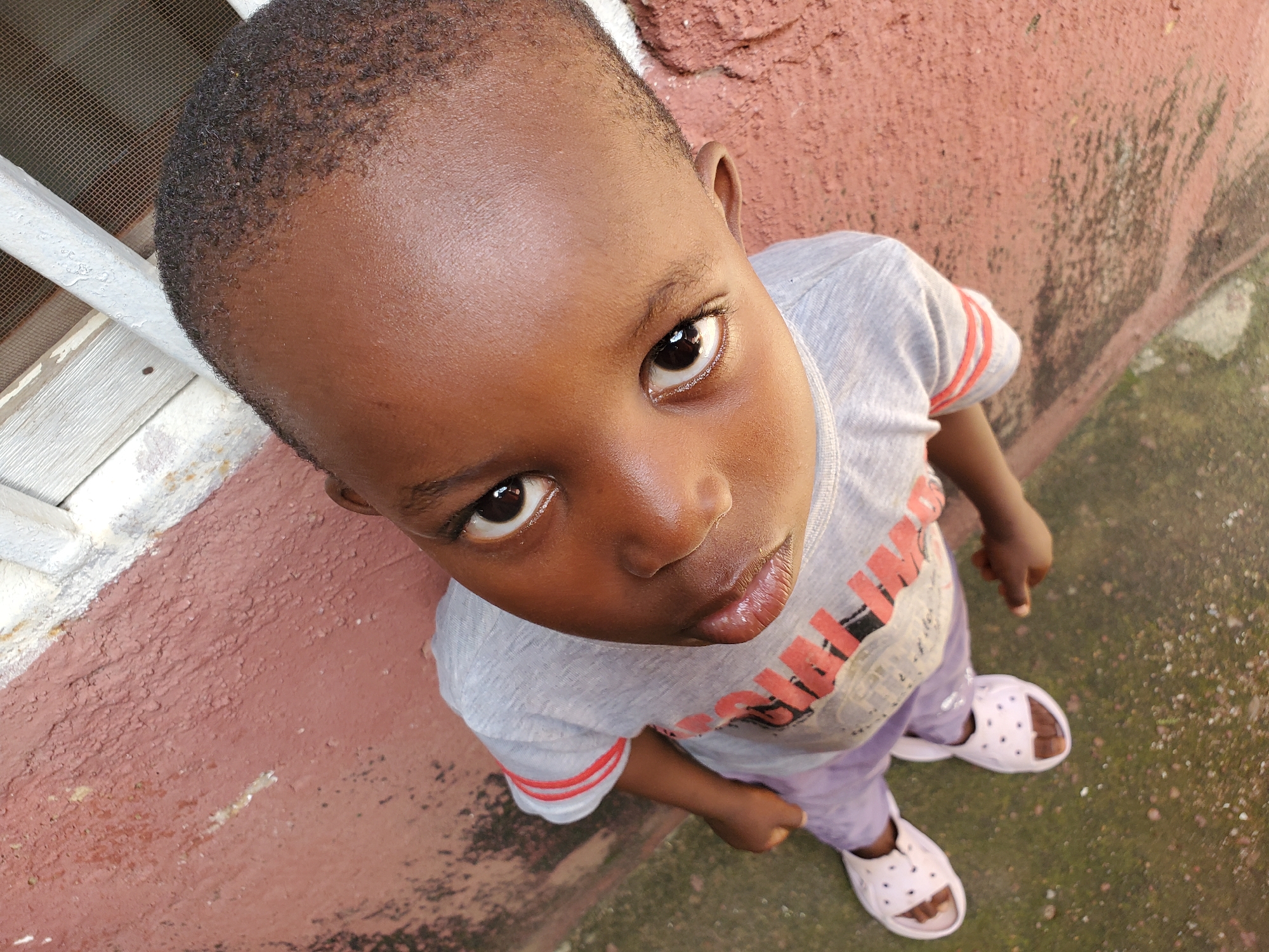 This is a high-angle, close-up shot of a young Black child, likely a toddler, standing outdoors. The child is wearing a light gray t-shirt with red and white striped sleeves and an orange graphic print on the front. The t-shirt has visible text that appears to be "AMAZING." The child is also wearing light purple pants and white slide sandals with a dotted pattern.
The child's skin tone is dark brown, and they have short, dark, curly hair. Their eyes are dark brown, and their lips are slightly parted. The child's expression appears curious and perhaps a little serious as they look up towards the camera.
The background consists of a textured, reddish-brown wall on the left side of the frame and a mottled green and brown ground, possibly concrete or pavement with some moss or dirt, on the right. A white window frame with a wire mesh screen is visible on the far left. The lighting suggests it is daytime, possibly in the late afternoon given the warmth of the light. The overall scene appears to be an outdoor area in a residential or public space. The location context is Jos, Nigeria.