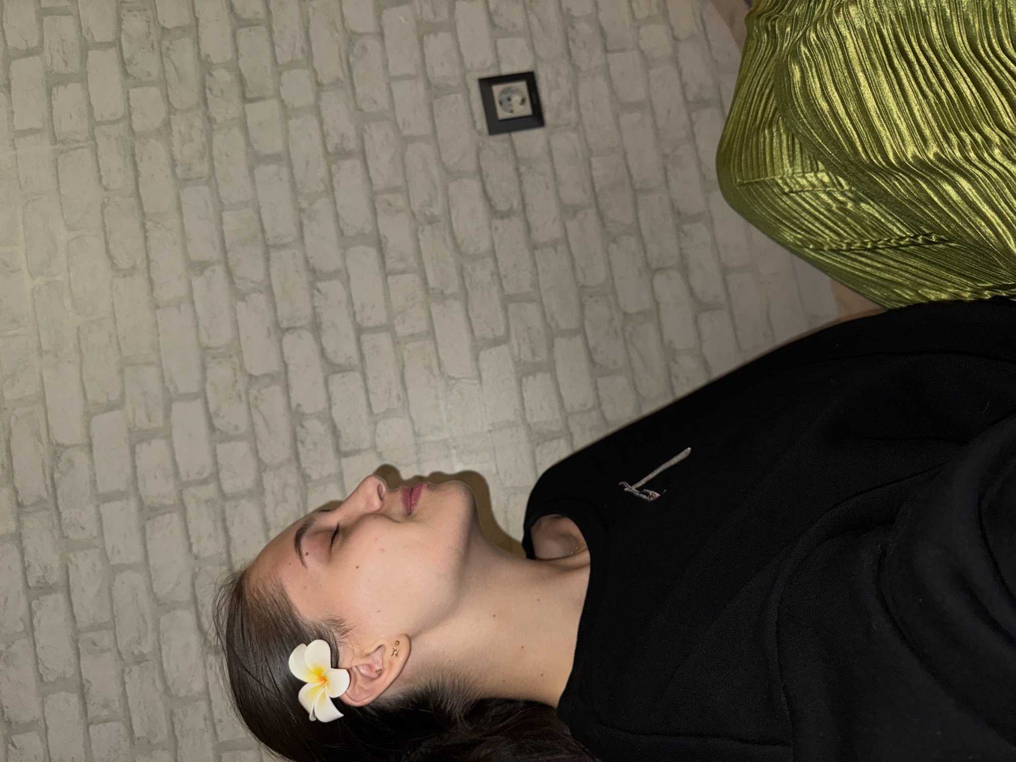 The image shows a young woman from the side, with her eyes closed and a flower tucked behind her ear. She is wearing a black top and a shiny green skirt. She has dark hair pulled back and small earrings in her ears. Behind her is a brick wall. There is an electrical outlet on the wall to the right of the woman. The overall lighting is dim.
This image is not in Sofia, Bulgaria.