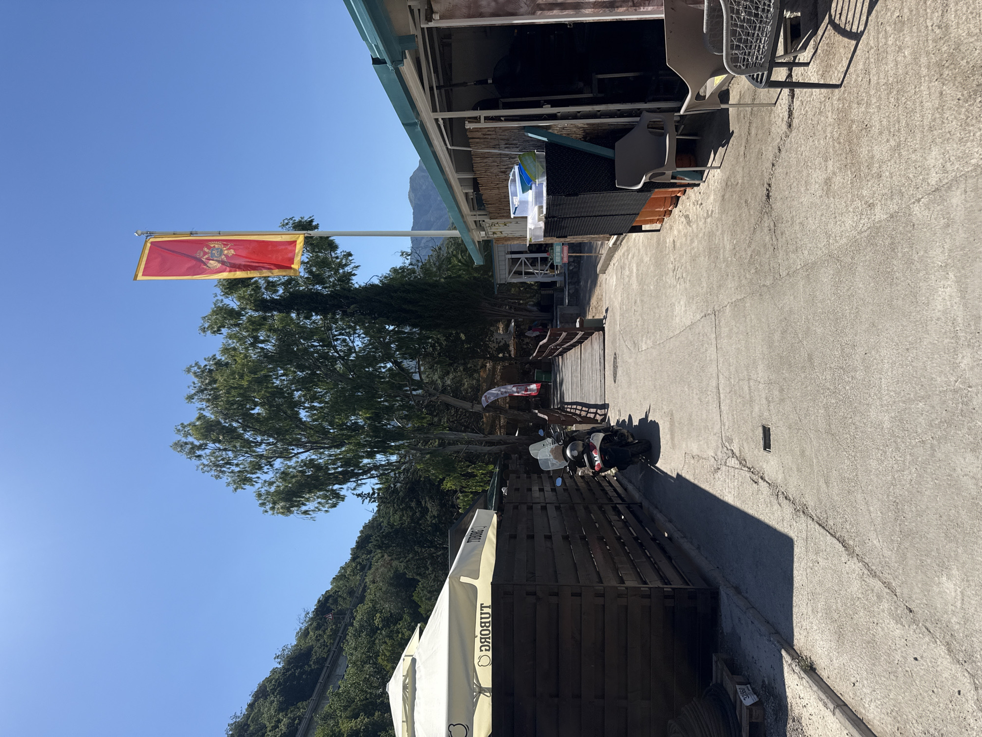The image depicts an outdoor scene on a sunny day, likely in Kamenovo, Montenegro. A flag of Montenegro, red with a golden border and its coat of arms, flutters from a pole against a clear blue sky. The setting appears to be an outdoor seating area or walkway adjacent to a building, possibly a restaurant or cafe. In the background, lush green trees and a distant mountain are visible, suggesting a coastal or hilly location.
Several objects are present, including outdoor furniture such as chairs and tables, some with a woven texture and others with a solid grey design. A scooter is parked on the right side of the walkway. A large white umbrella or awning with visible text on it extends from the left side of the frame, partially obscuring a wooden fence structure. The ground is paved with concrete, showing some cracks and casting distinct shadows.
The scene suggests a relaxed, possibly tourist-oriented environment. The bright sunlight and clear sky indicate it is daytime, likely mid-morning to afternoon. There are no people clearly identifiable in the foreground, but the presence of seating and a scooter implies recent or potential human activity. The overall impression is one of a pleasant, sunny day in a scenic location. Text visible on the umbrella is "LUBOBG", which appears to be a brand name or logo.
