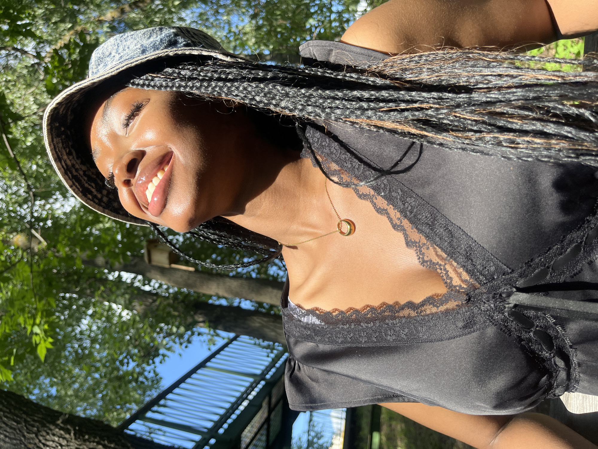 A young Black woman with braided hair and a bucket hat is captured in a close-up, outdoor shot. She is looking upwards with her eyes closed and a wide smile, her face illuminated by the sun. She is wearing a black lace-trimmed top and a gold necklace with a circular pendant. The background shows lush green trees, a blue sky, and a glimpse of a dark railing structure, possibly a bridge or walkway. The scene appears to be set in a park or natural area. There is no visible indication of the location being the city of Kazan, Russia.