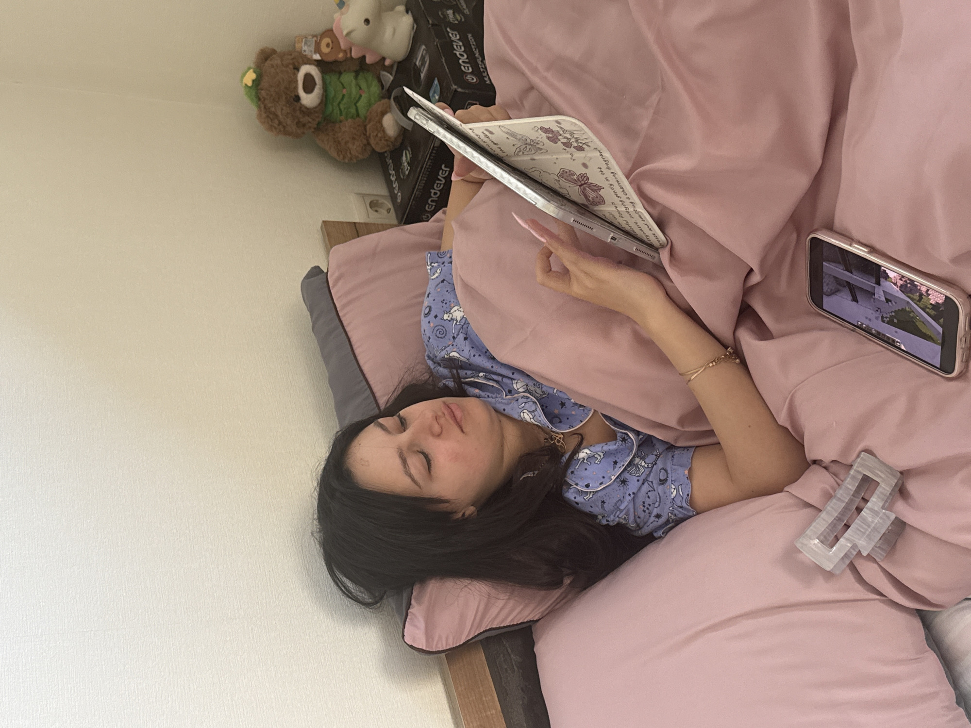 A woman with dark hair and wearing blue pajamas is lying in a bed with pink sheets. She is holding a white tablet in her right hand. A smartphone sits on the bed to her right, displaying a video. To the left of the phone is a hair clip. Behind her are a teddy bear and a box labeled "Endever." The background appears to be a white wall. The location is unknown and cannot be inferred from the image.