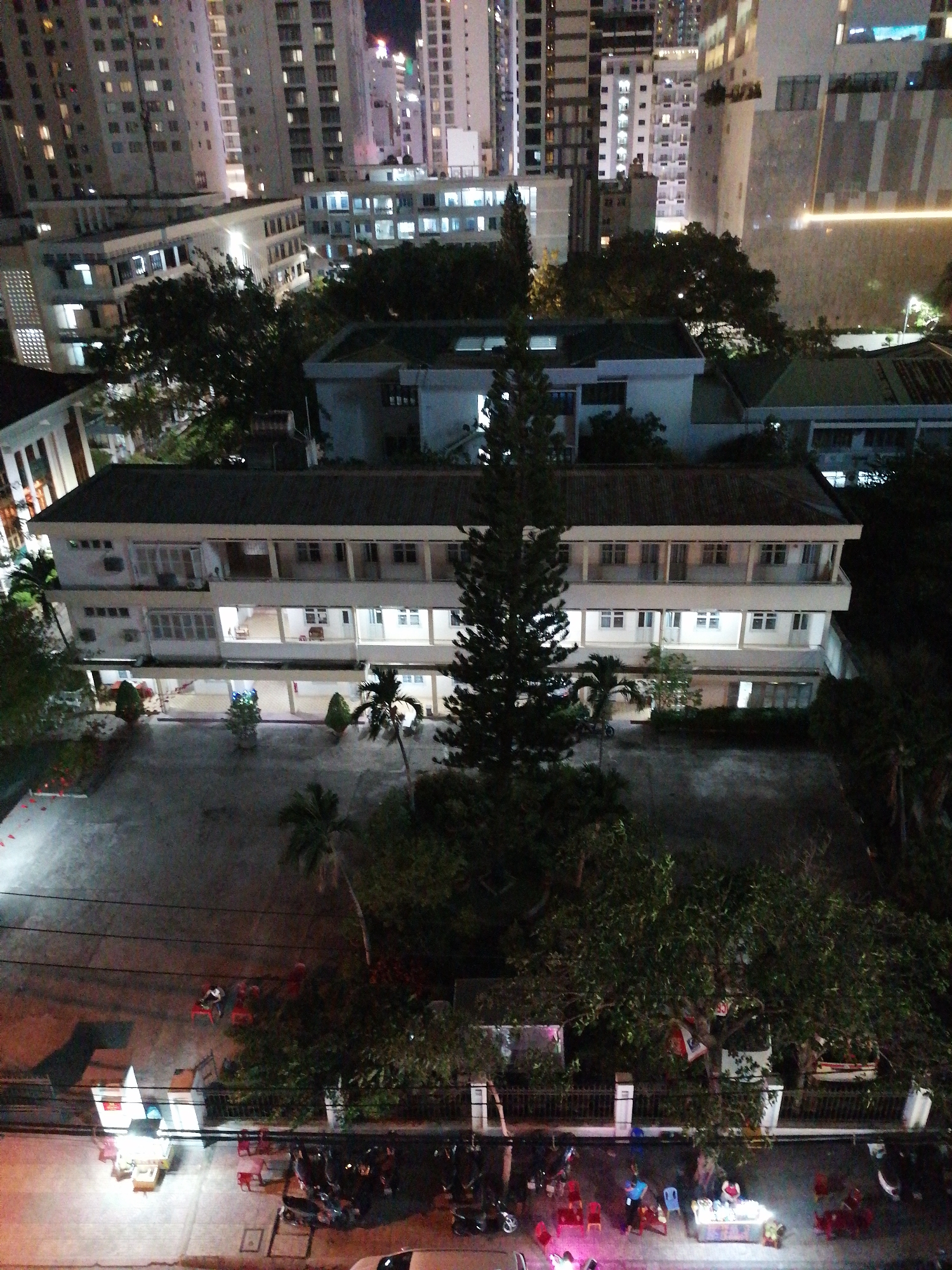 An aerial shot in Nha Trang Ward, Vietnam, shows a two-story white building with a dark roof, centered in the frame. The building features a balcony on both floors. A tall, dark evergreen tree stands prominently in front of the building, obscuring part of its facade. A paved courtyard stretches in front of the building, sparsely populated with small trees and plants. Beyond the courtyard, a fence separates the building from a street scene. On the street, several motorbikes are parked. A brightly lit food stall is set up, surrounded by small red chairs and stools. People are visible near the stall. The background is dominated by several high-rise buildings, their windows illuminated at night. The overall lighting is a mix of artificial lights from the buildings and street.